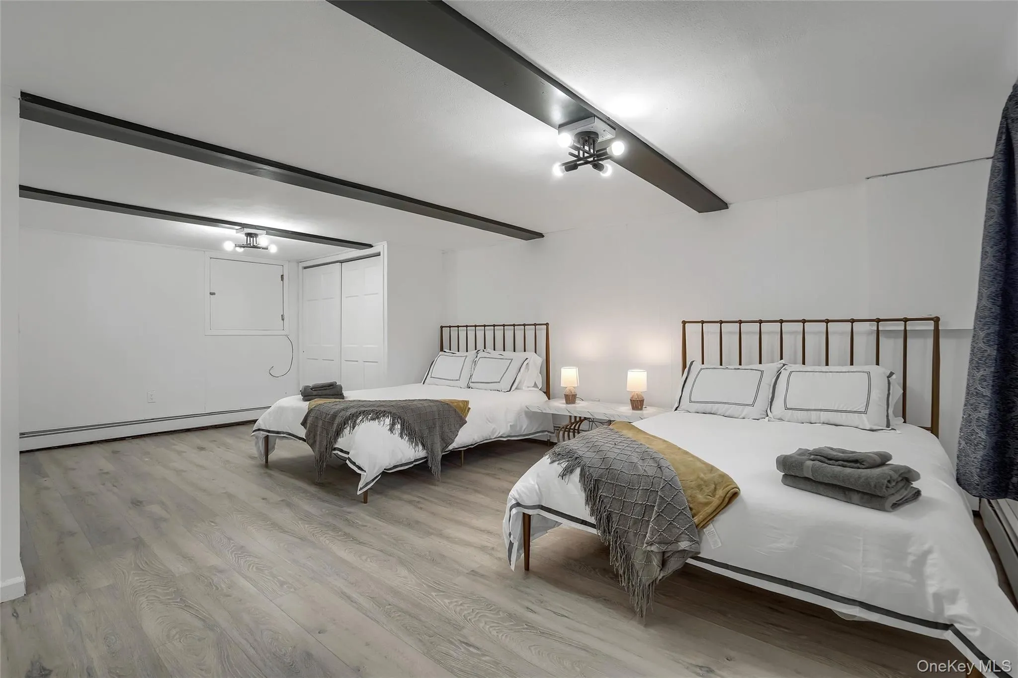 Spacious room featuring light wood-finish flooring, white walls, exposed ceiling beams, and dual overhead light fixtures Spacious room featuring light wood-finish flooring, white walls, exposed ceiling beams, and dual overhead light fixtures