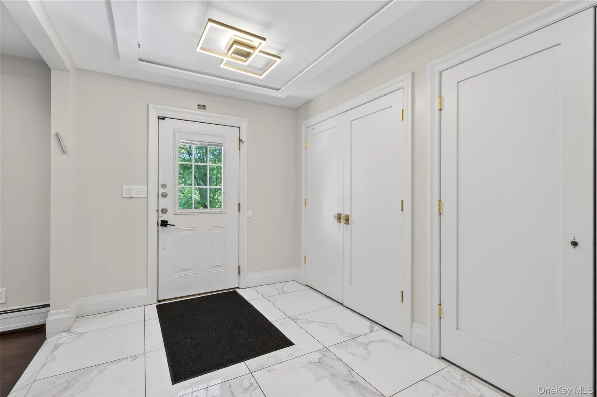 Foyer featuring white marble-look tile flooring, a recessed ceiling with a contemporary light fixture, and a white entry door with a window pane Foyer featuring white marble-look tile flooring, a recessed ceiling with a contemporary light fixture, and a white entry door with a window pane
