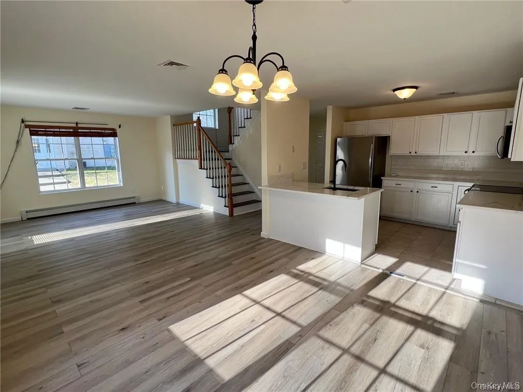 Kitchen with white cabinetry, light wood-type flooring, stainless steel appliances, a notable chandelier, and baseboard heating and this area also shows the dining area and living room. Kitchen with white cabinetry, light wood-type flooring, stainless steel appliances, a notable chandelier, and baseboard heating and this area also shows the dining area and living room.