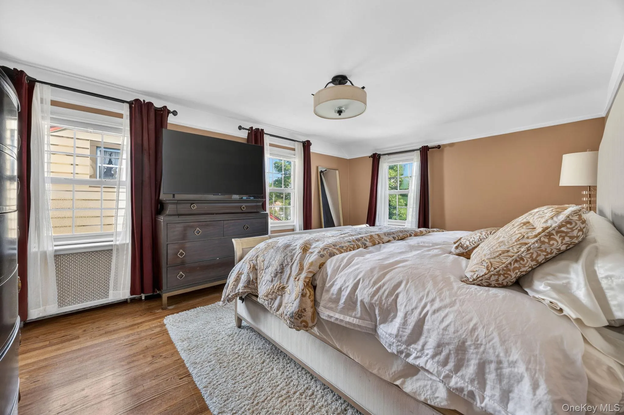 Spacious room featuring wood-finish flooring, multiple windows, a ceiling-mounted light fixture, crown molding, and a neutral color palette Spacious room featuring wood-finish flooring, multiple windows, a ceiling-mounted light fixture, crown molding, and a neutral color palette