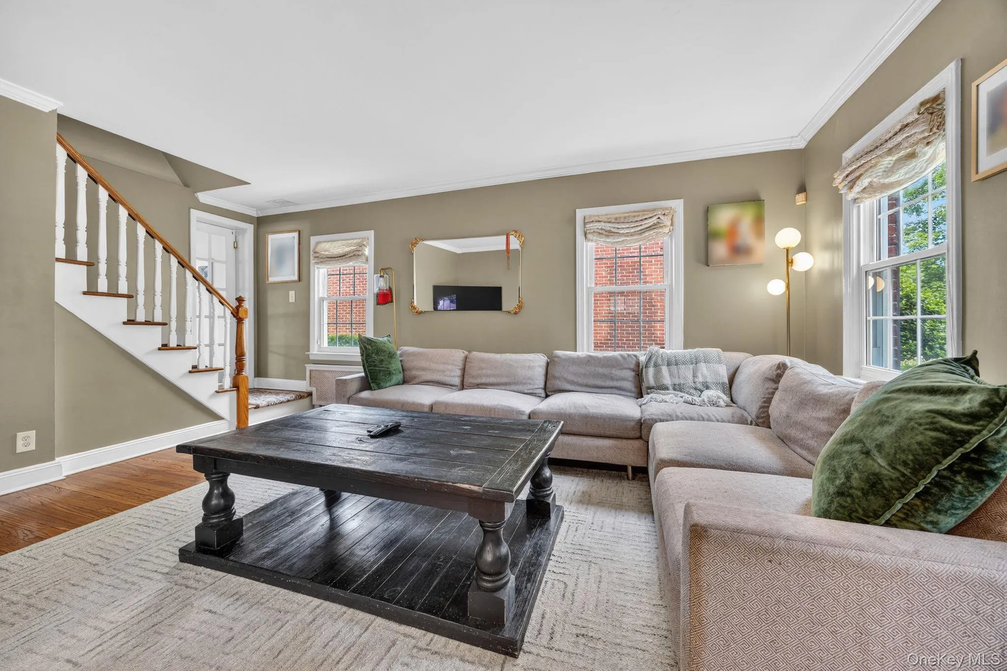 Living area featuring hardwood flooring, a white staircase with wooden handrail, crown molding, and multiple windows providing natural light Living area featuring hardwood flooring, a white staircase with wooden handrail, crown molding, and multiple windows providing natural light
