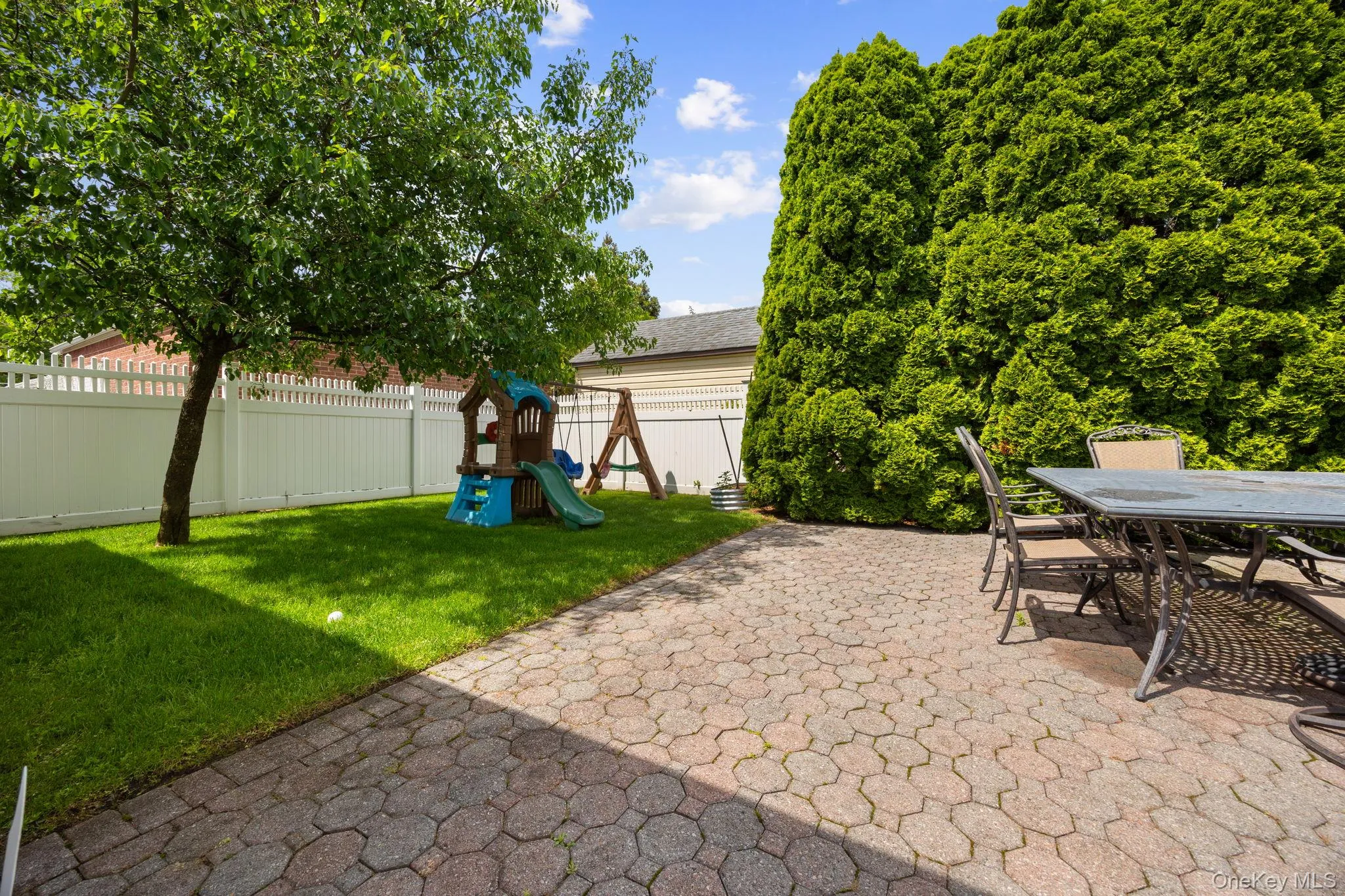 Paved patio area featuring hexagonal pavers, bordering a grass lawn with a mature trees, and a white fence Paved patio area featuring hexagonal pavers, bordering a grass lawn with a mature trees, and a white fence