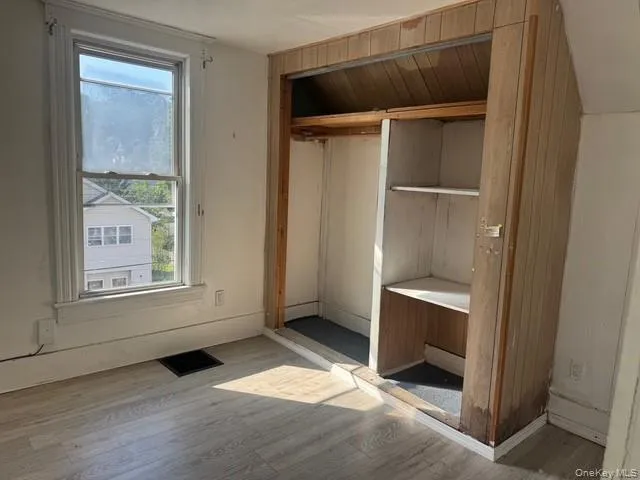 Room featuring wood-finish flooring, a double-hung window, and a built-in closet with shelving and wood paneling Room featuring wood-finish flooring, a double-hung window, and a built-in closet with shelving and wood paneling
