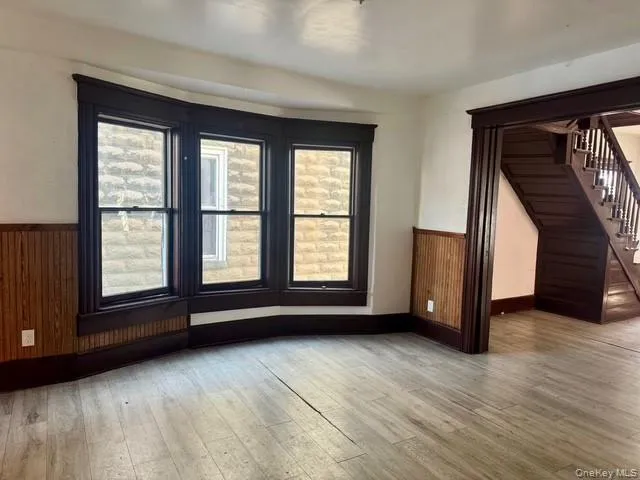 Living area featuring light wood-finish flooring, dark wood trim, and a three-panel bay window Living area featuring light wood-finish flooring, dark wood trim, and a three-panel bay window