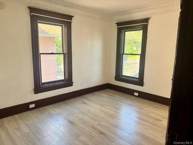 Room featuring two single-hung windows with dark wood-finish trim, light-toned wood-finish flooring, white walls, and decorative crown molding Room featuring two single-hung windows with dark wood-finish trim, light-toned wood-finish flooring, white walls, and decorative crown molding