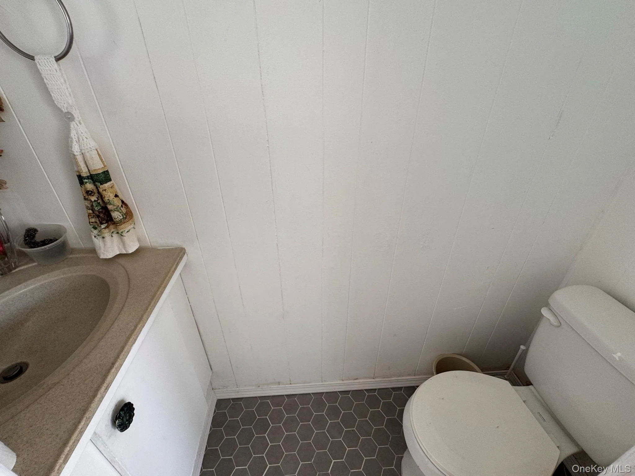Compact bathroom featuring hexagonal floor tiling, white paneled walls, a built-in vanity with an integrated sink, and a commode Compact bathroom featuring hexagonal floor tiling, white paneled walls, a built-in vanity with an integrated sink, and a commode