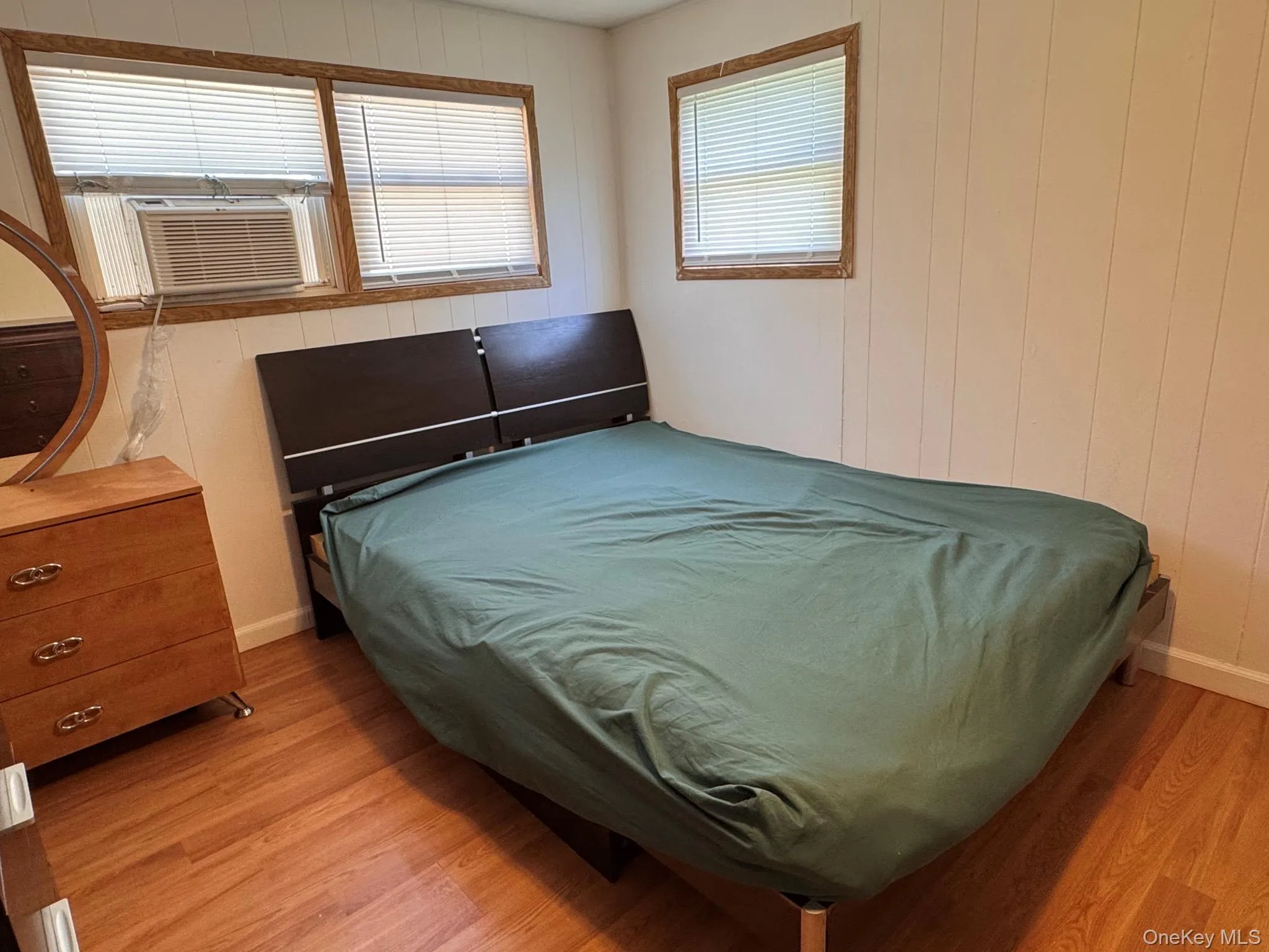 Room featuring light-toned vertical paneling, wood-finish flooring, and multiple windows with blinds Room featuring light-toned vertical paneling, wood-finish flooring, and multiple windows with blinds