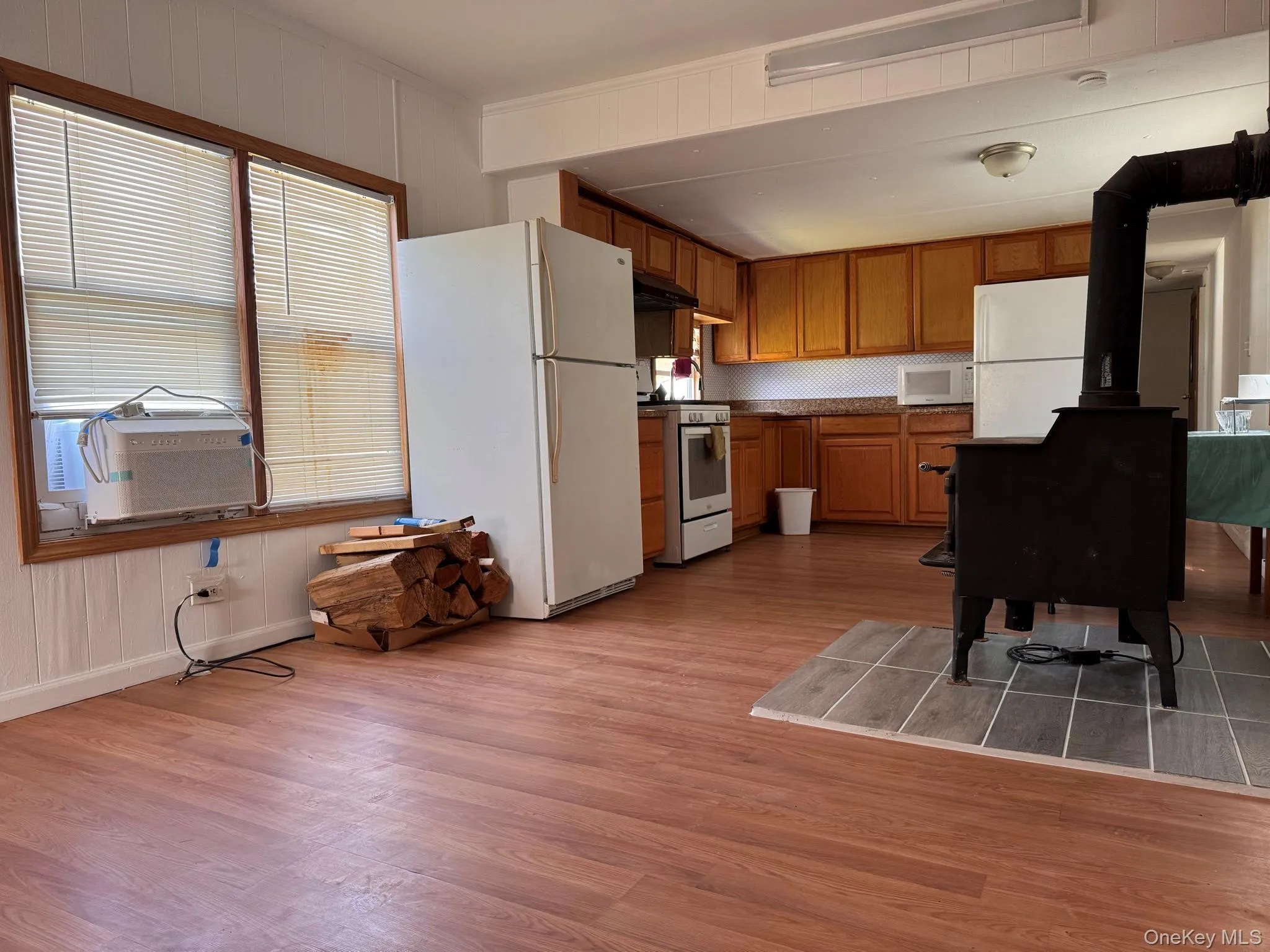 Open-concept living area featuring wood-finish flooring, a wood-burning stove with a tile hearth, and a functional kitchen space Open-concept living area featuring wood-finish flooring, a wood-burning stove with a tile hearth, and a functional kitchen space