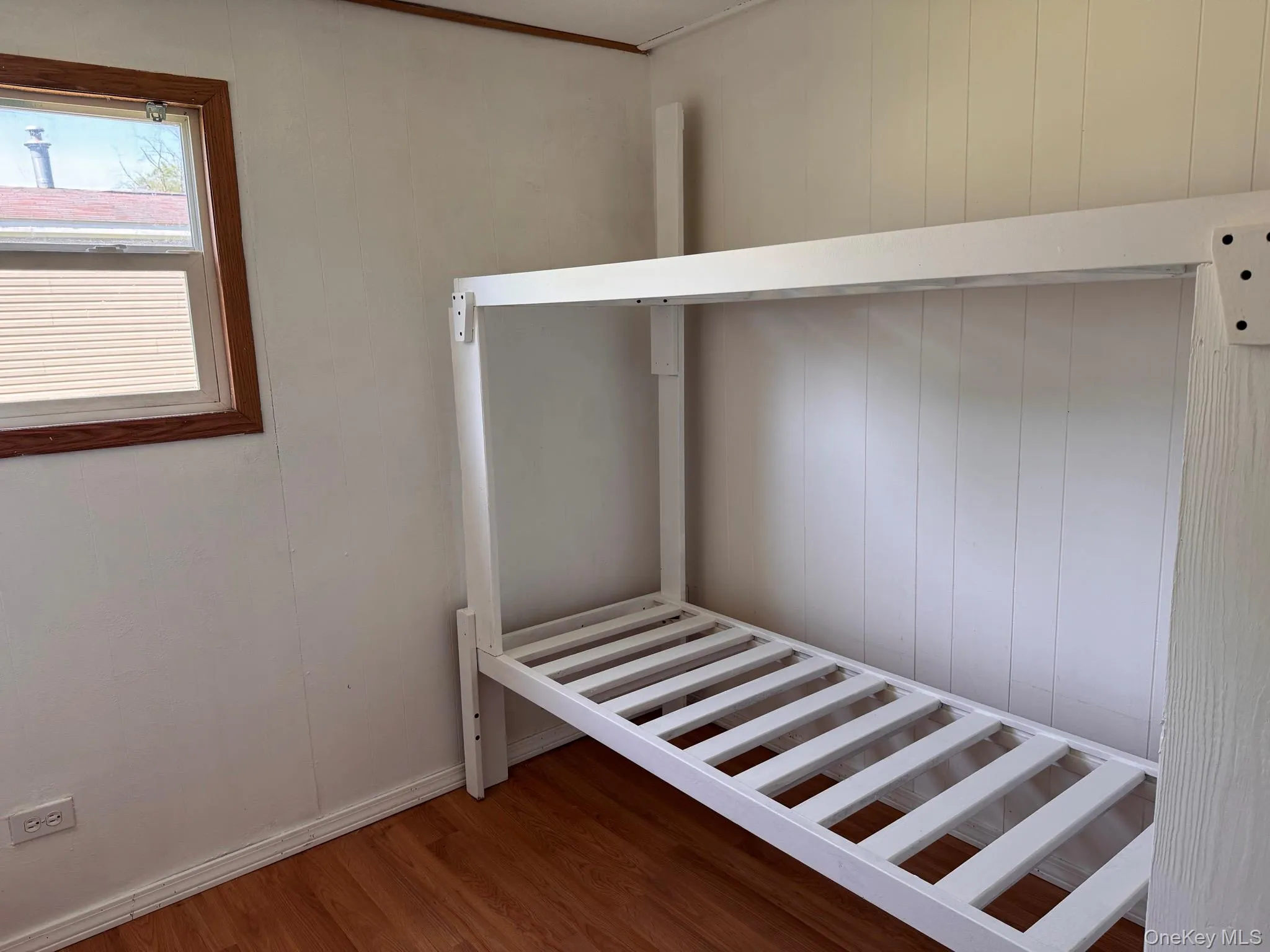 Interior room featuring wood-finish flooring, white paneling, and a single window with wood trim Interior room featuring wood-finish flooring, white paneling, and a single window with wood trim