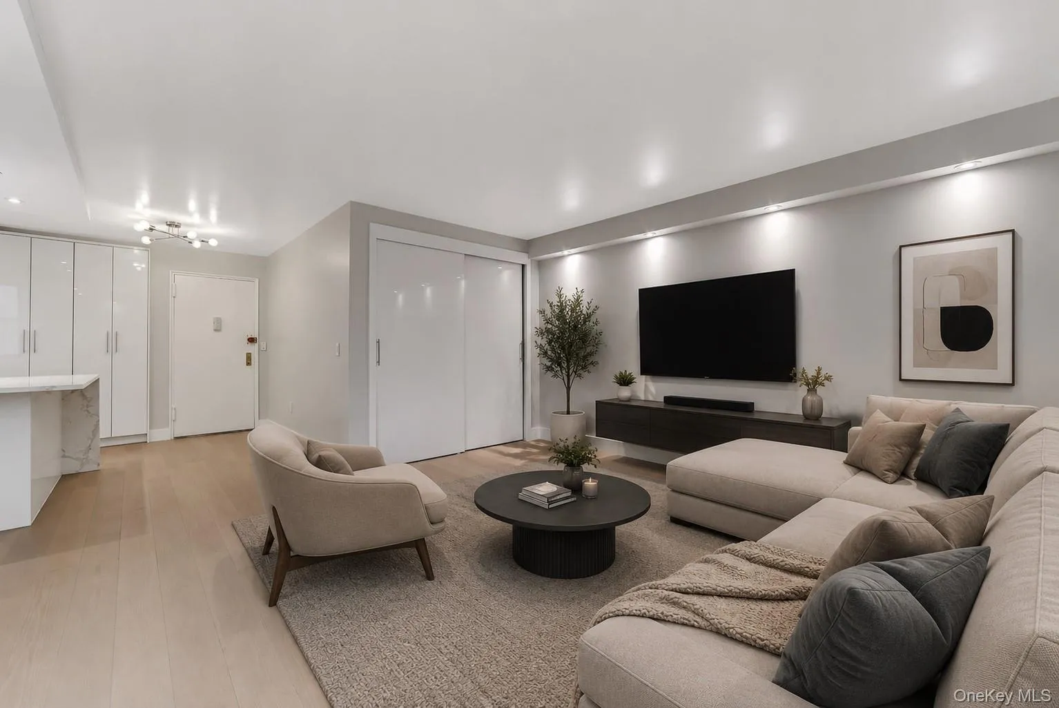 Open-concept living area featuring light wood-finish flooring, a wall-mounted media console, recessed lighting, and a built-in storage unit with a marble-effect countertop Open-concept living area featuring light wood-finish flooring, a wall-mounted media console, recessed lighting, and a built-in storage unit with a marble-effect countertop