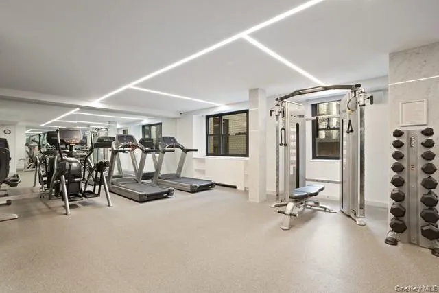 Fitness center featuring recessed linear lighting, mirrored wall panels, multiple cardio machines, a cable cross machine, and a dumbbell rack Fitness center featuring recessed linear lighting, mirrored wall panels, multiple cardio machines, a cable cross machine, and a dumbbell rack