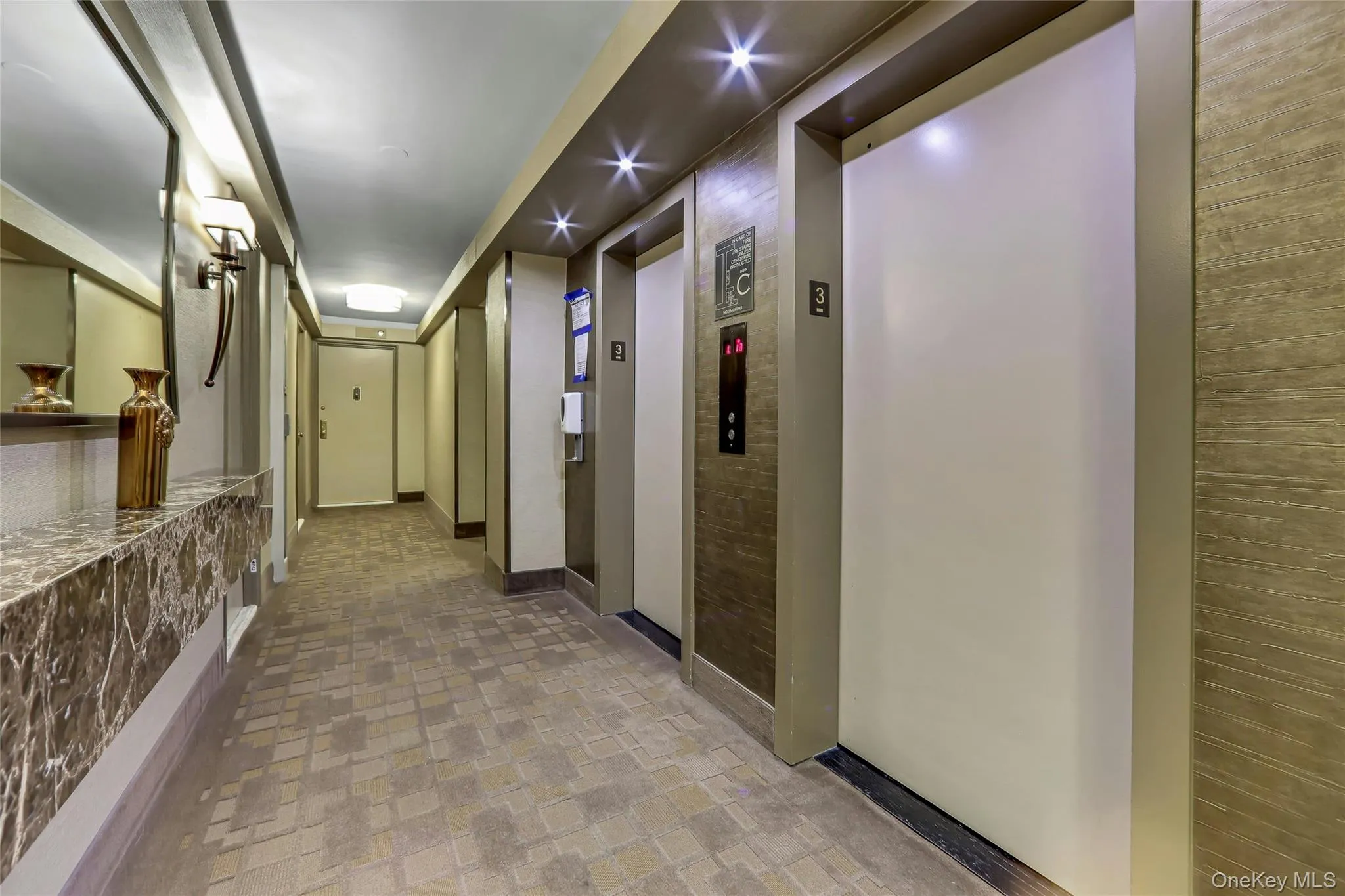 Elevator lobby featuring two elevator banks, recessed ceiling lighting, a decorative wall sconce, and a built-in marble-look console table Elevator lobby featuring two elevator banks, recessed ceiling lighting, a decorative wall sconce, and a built-in marble-look console table
