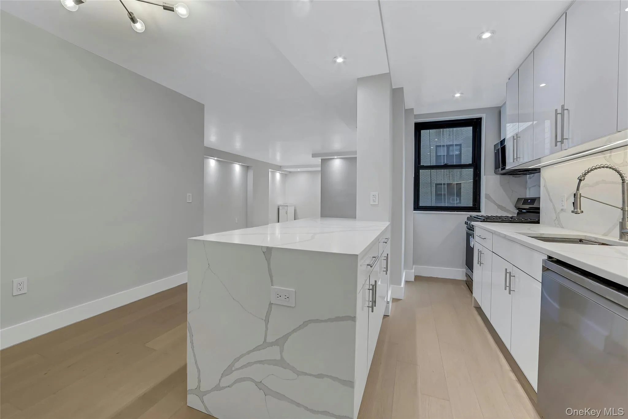 Open-concept kitchen featuring a waterfall island with stone countertop, white flat-panel cabinetry, stainless steel appliances, and wood-finish flooring Open-concept kitchen featuring a waterfall island with stone countertop, white flat-panel cabinetry, stainless steel appliances, and wood-finish flooring