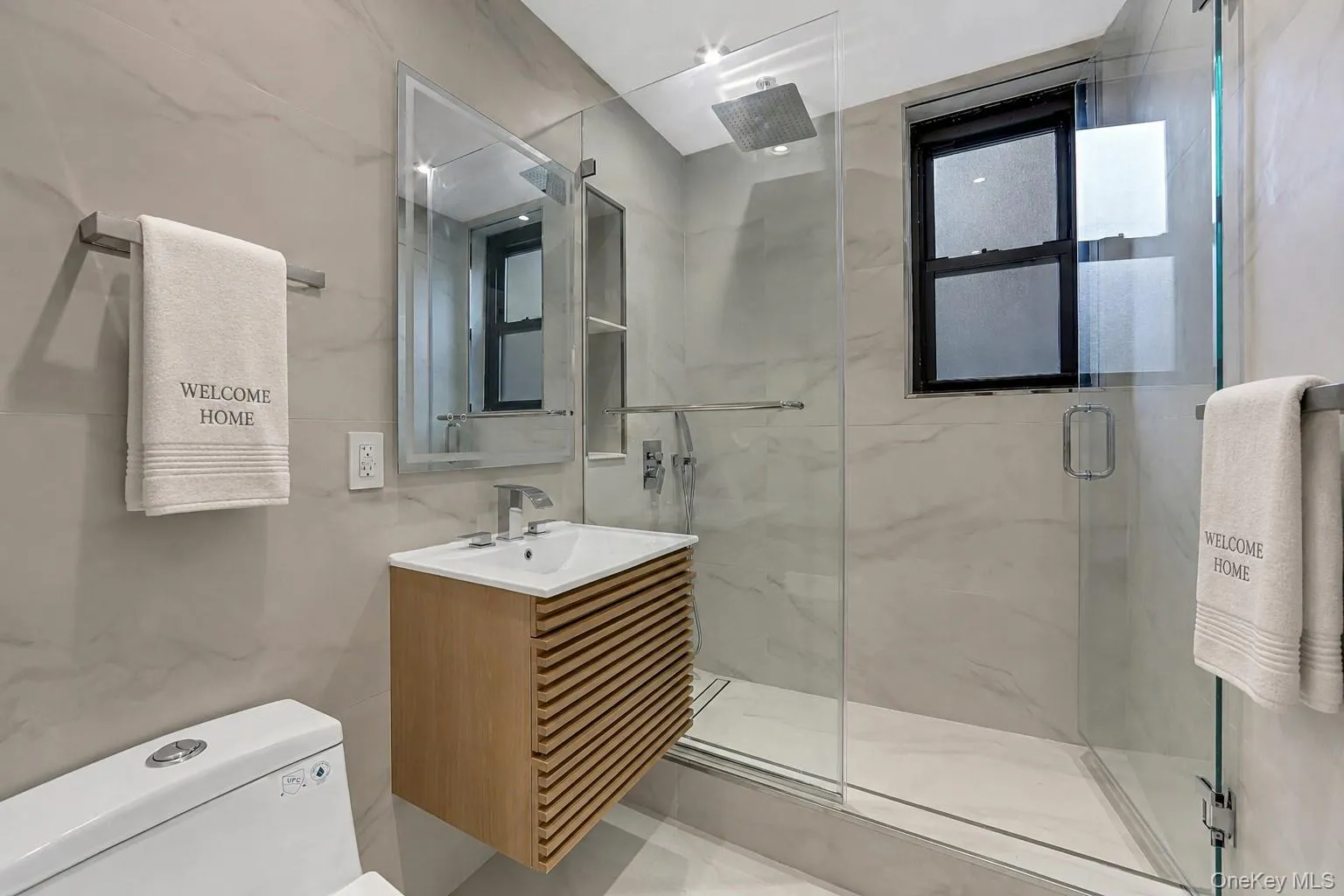 Modern bathroom featuring a glass-enclosed shower with a rainfall showerhead, a floating wood-finish vanity with an integrated sink, and large format marble-look tile walls Modern bathroom featuring a glass-enclosed shower with a rainfall showerhead, a floating wood-finish vanity with an integrated sink, and large format marble-look tile walls