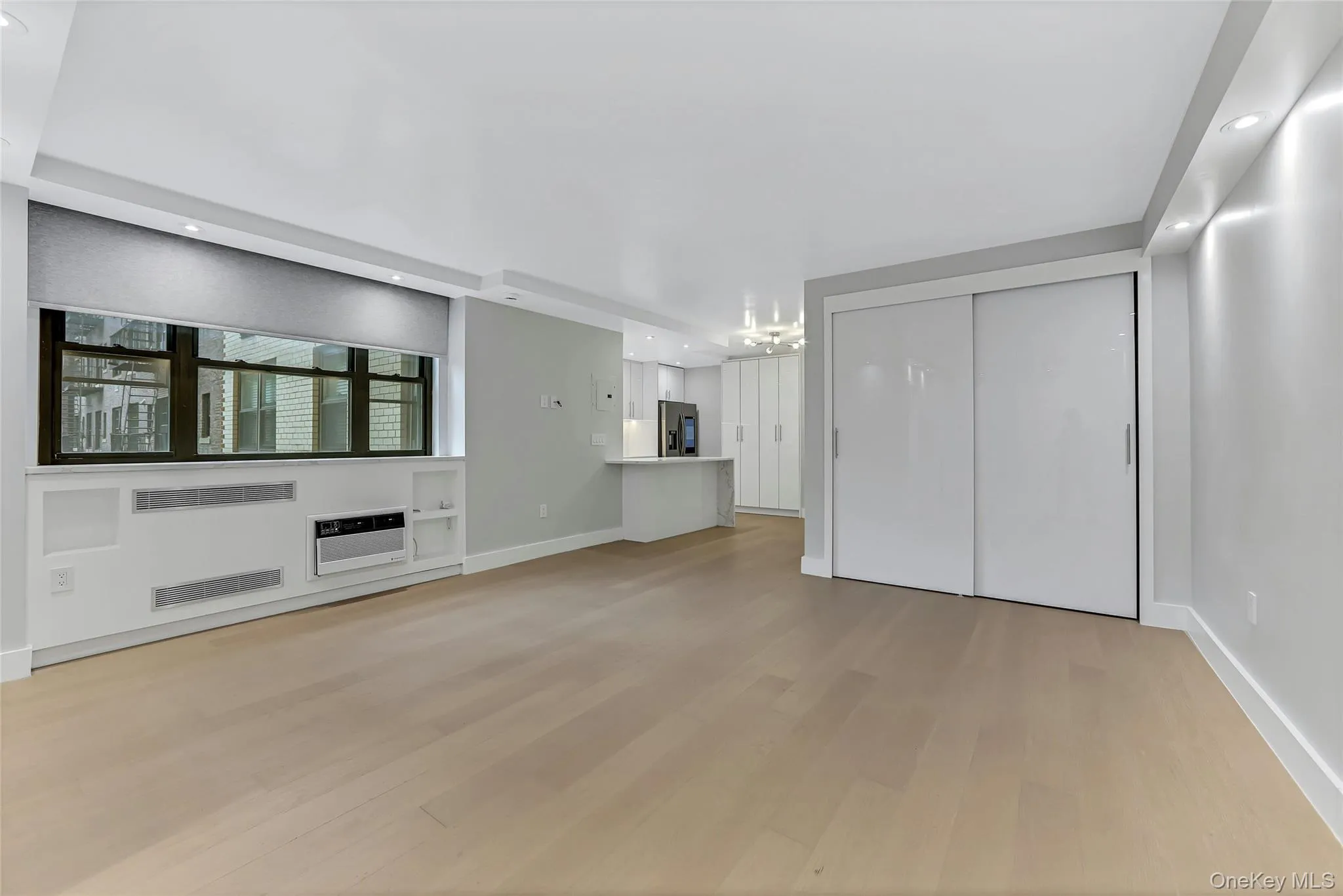 Open concept living area featuring light wood-finish flooring, recessed ceiling lighting, and a large window with roller shades Open concept living area featuring light wood-finish flooring, recessed ceiling lighting, and a large window with roller shades