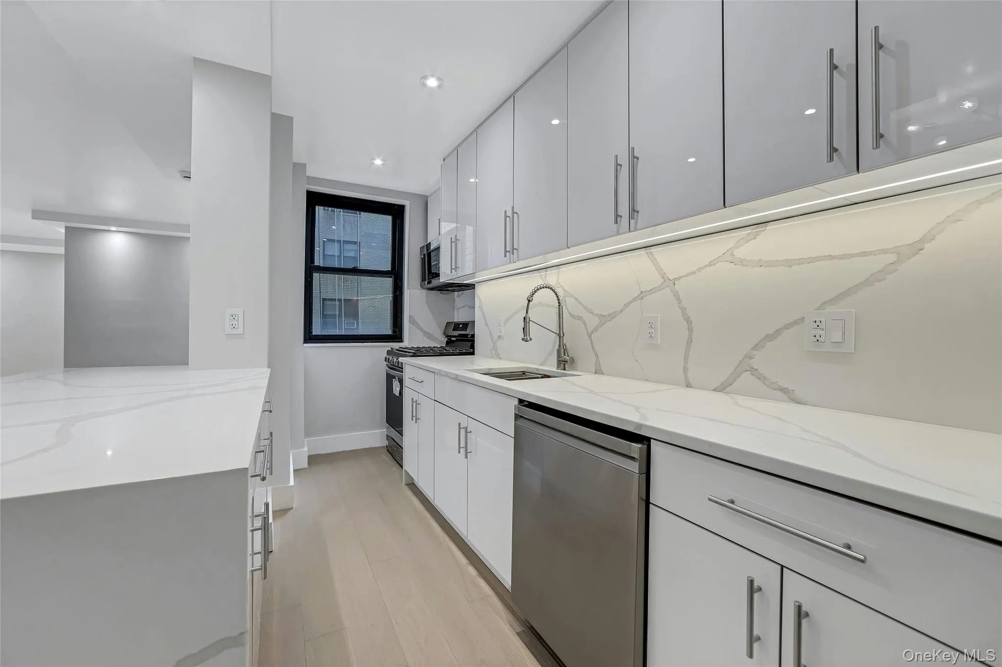 Modern kitchen featuring white cabinetry, integrated stainless steel appliances, light wood-finish flooring, white countertop surfaces, and a matching backsplash Modern kitchen featuring white cabinetry, integrated stainless steel appliances, light wood-finish flooring, white countertop surfaces, and a matching backsplash