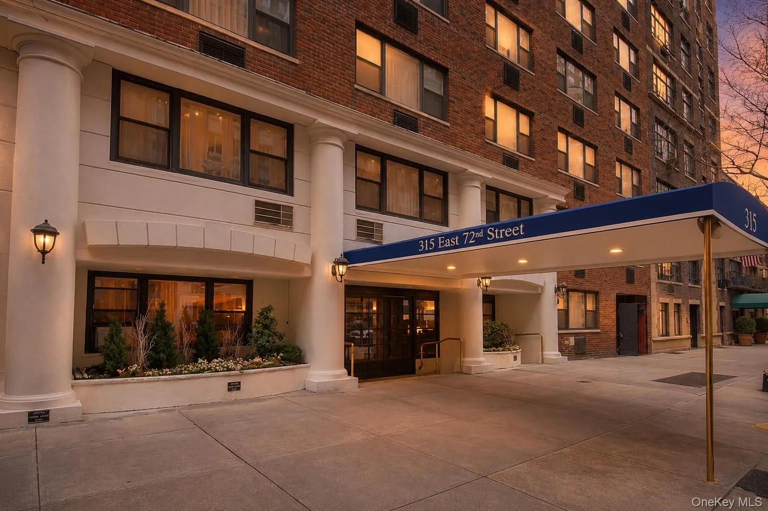 Brick building facade featuring a covered entrance with a blue awning, decorative outdoor lighting, large street-level windows, and a landscaped planter Brick building facade featuring a covered entrance with a blue awning, decorative outdoor lighting, large street-level windows, and a landscaped planter