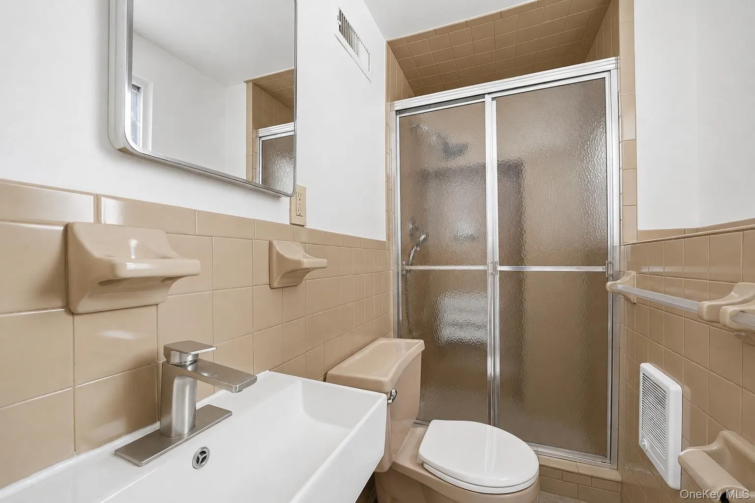 Basement bathroom featuring tan tiled walls, a white rectangular sink with a brushed nickel faucet, and a shower with frosted glass sliding doors Basement bathroom featuring tan tiled walls, a white rectangular sink with a brushed nickel faucet, and a shower with frosted glass sliding doors