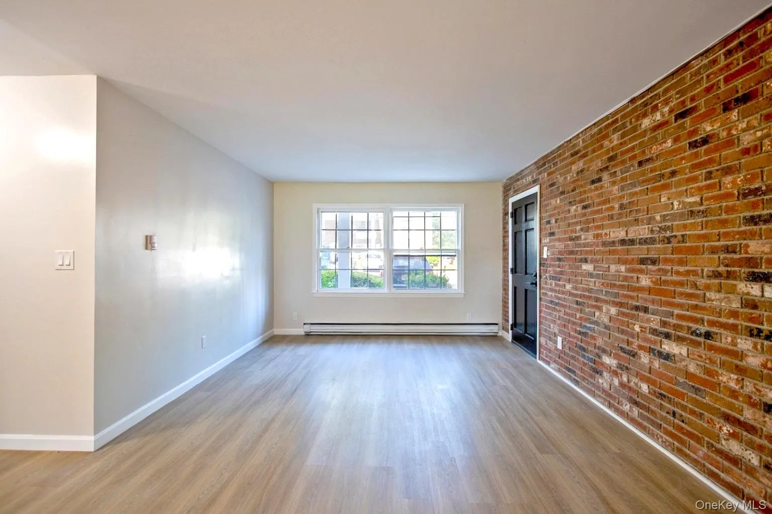 Spacious room featuring a prominent exposed brick accent wall, wood-finish flooring, and white baseboards Spacious room featuring a prominent exposed brick accent wall, wood-finish flooring, and white baseboards
