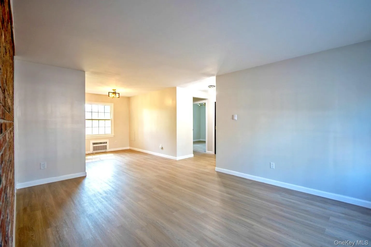 Spacious interior featuring wood-finish flooring, light-colored walls, exposed brick accent wall, and a window with a room air conditioning unit Spacious interior featuring wood-finish flooring, light-colored walls, exposed brick accent wall, and a window with a room air conditioning unit