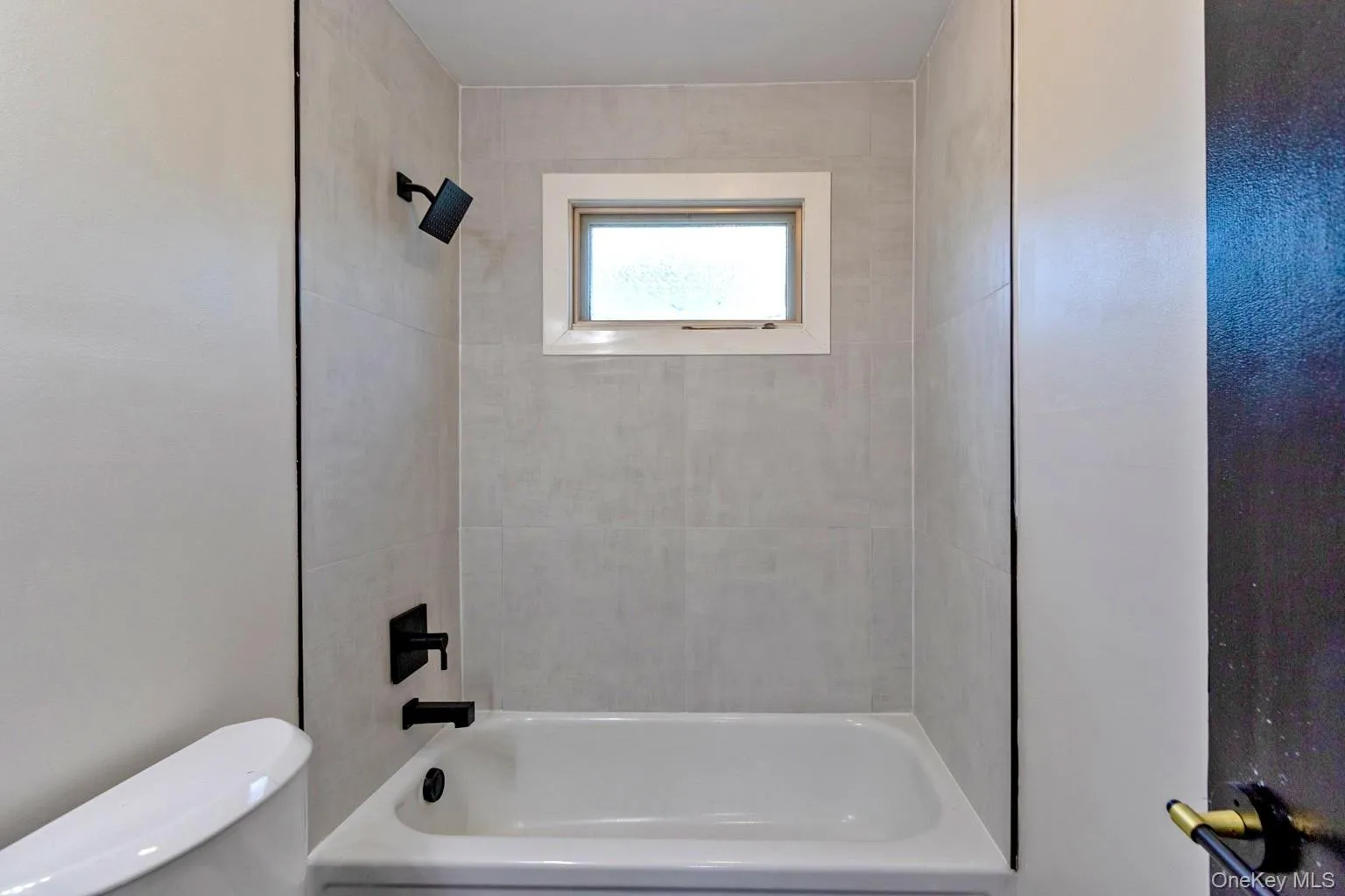 Shower and tub combination featuring light-toned large format tiling, a rectangular window with white trim, and matte black fixtures Shower and tub combination featuring light-toned large format tiling, a rectangular window with white trim, and matte black fixtures
