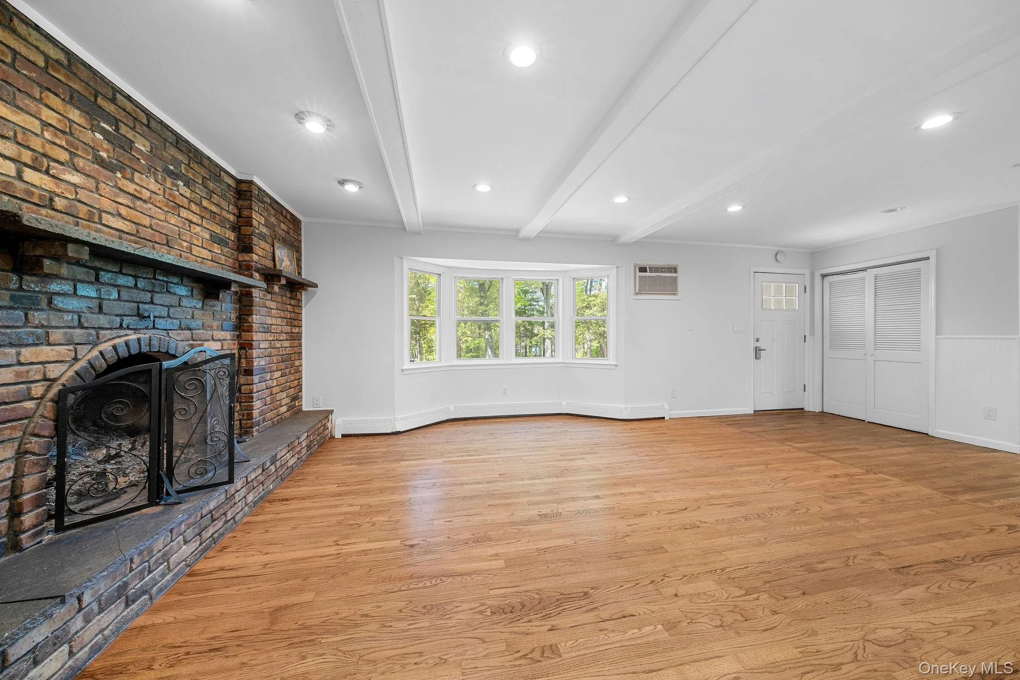 Spacious room featuring wood-finish flooring, a brick fireplace with hearth, a bay window, recessed lighting, and exposed ceiling beams Spacious room featuring wood-finish flooring, a brick fireplace with hearth, a bay window, recessed lighting, and exposed ceiling beams