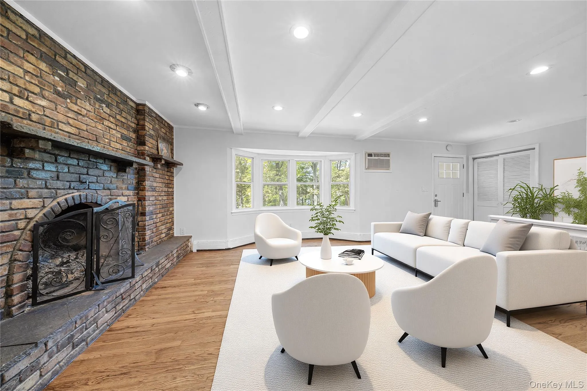 Spacious living area featuring a brick fireplace with a stone hearth, wood-finish flooring, a bay window, and exposed ceiling beams. Image is Virtually Staged. Spacious living area featuring a brick fireplace with a stone hearth, wood-finish flooring, a bay window, and exposed ceiling beams. Image is Virtually Staged.