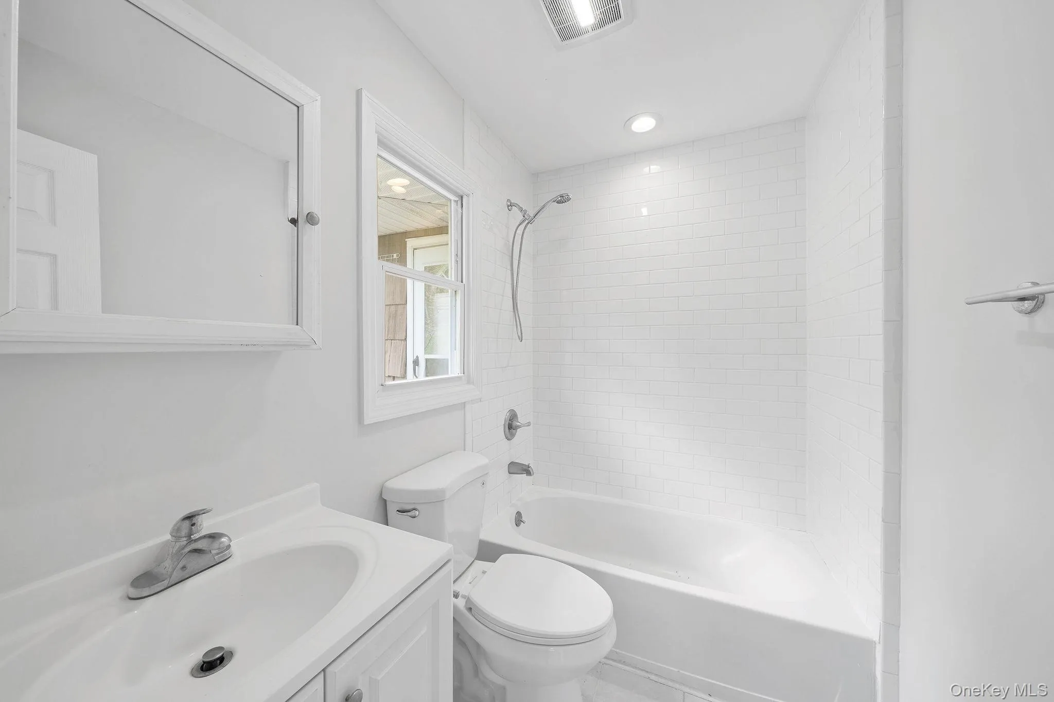 Bathroom featuring a white subway tile shower surround, a single-pane window, a white vanity with a single basin sink, a wall-mounted mirror, and a recessed ceiling light Bathroom featuring a white subway tile shower surround, a single-pane window, a white vanity with a single basin sink, a wall-mounted mirror, and a recessed ceiling light