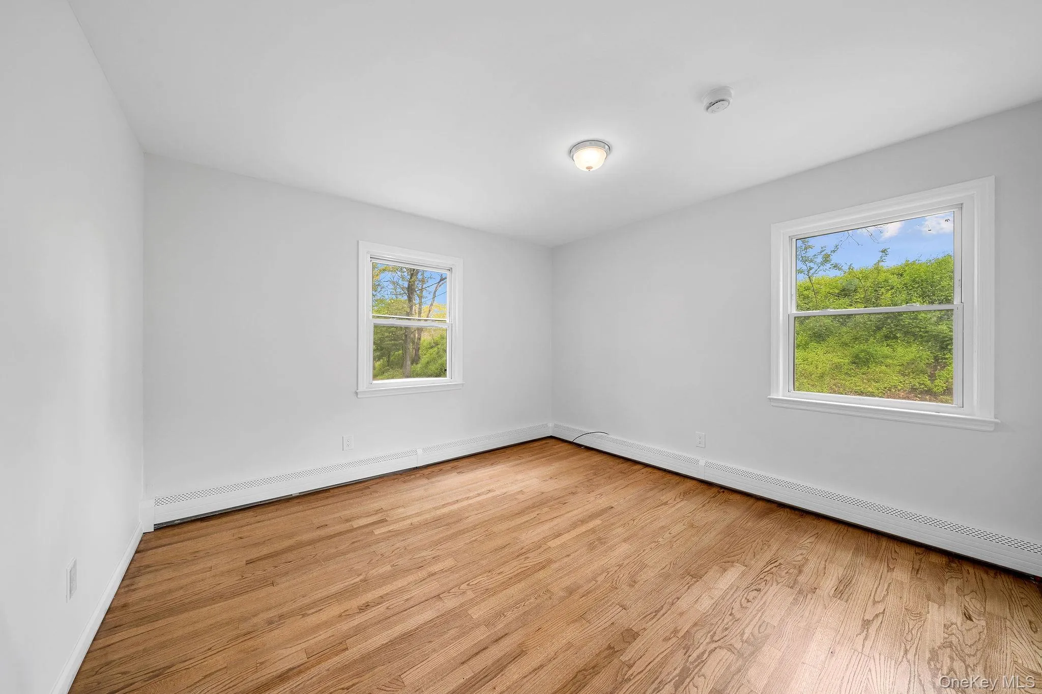 Hardwood flooring throughout a bright room featuring two windows with outdoor scenery Hardwood flooring throughout a bright room featuring two windows with outdoor scenery