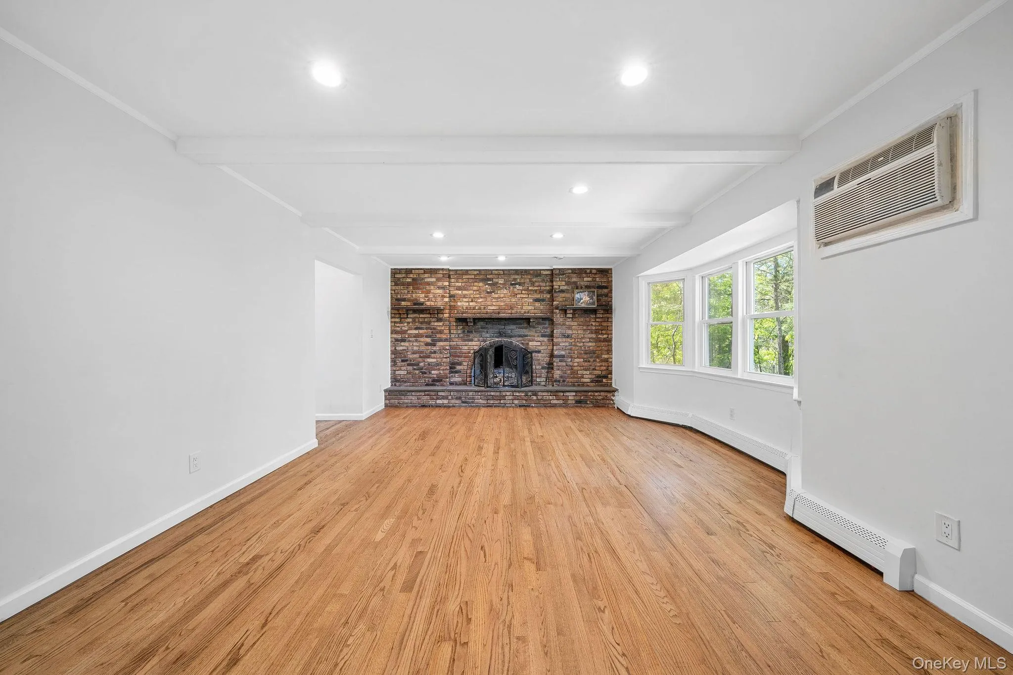 Living area featuring wood-finish flooring, a brick fireplace, a curved bay window, recessed lighting, and exposed ceiling beams Living area featuring wood-finish flooring, a brick fireplace, a curved bay window, recessed lighting, and exposed ceiling beams