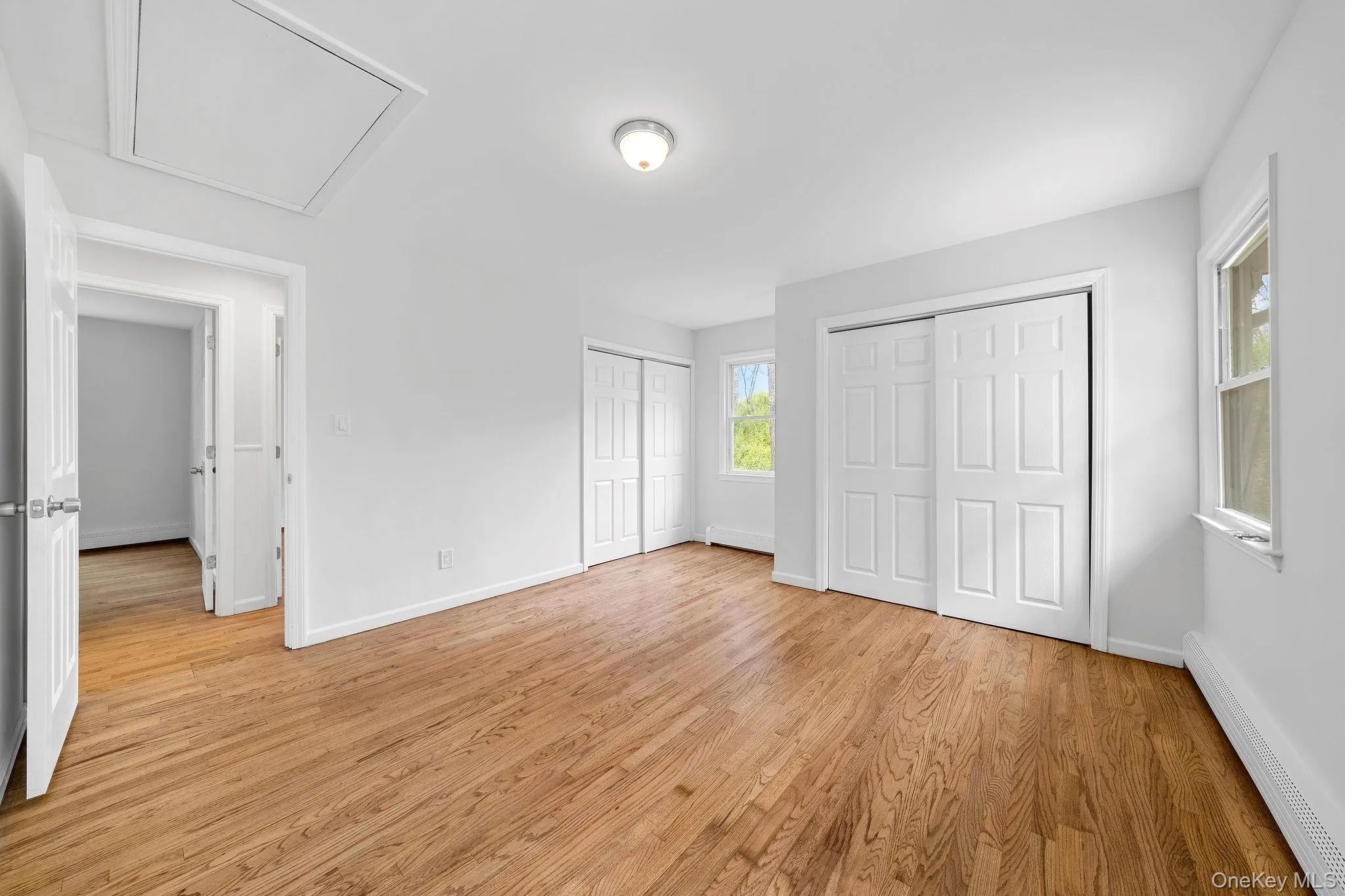 Spacious room featuring wood-finish flooring, white walls, multiple windows, and two sets of sliding panel closet doors Spacious room featuring wood-finish flooring, white walls, multiple windows, and two sets of sliding panel closet doors