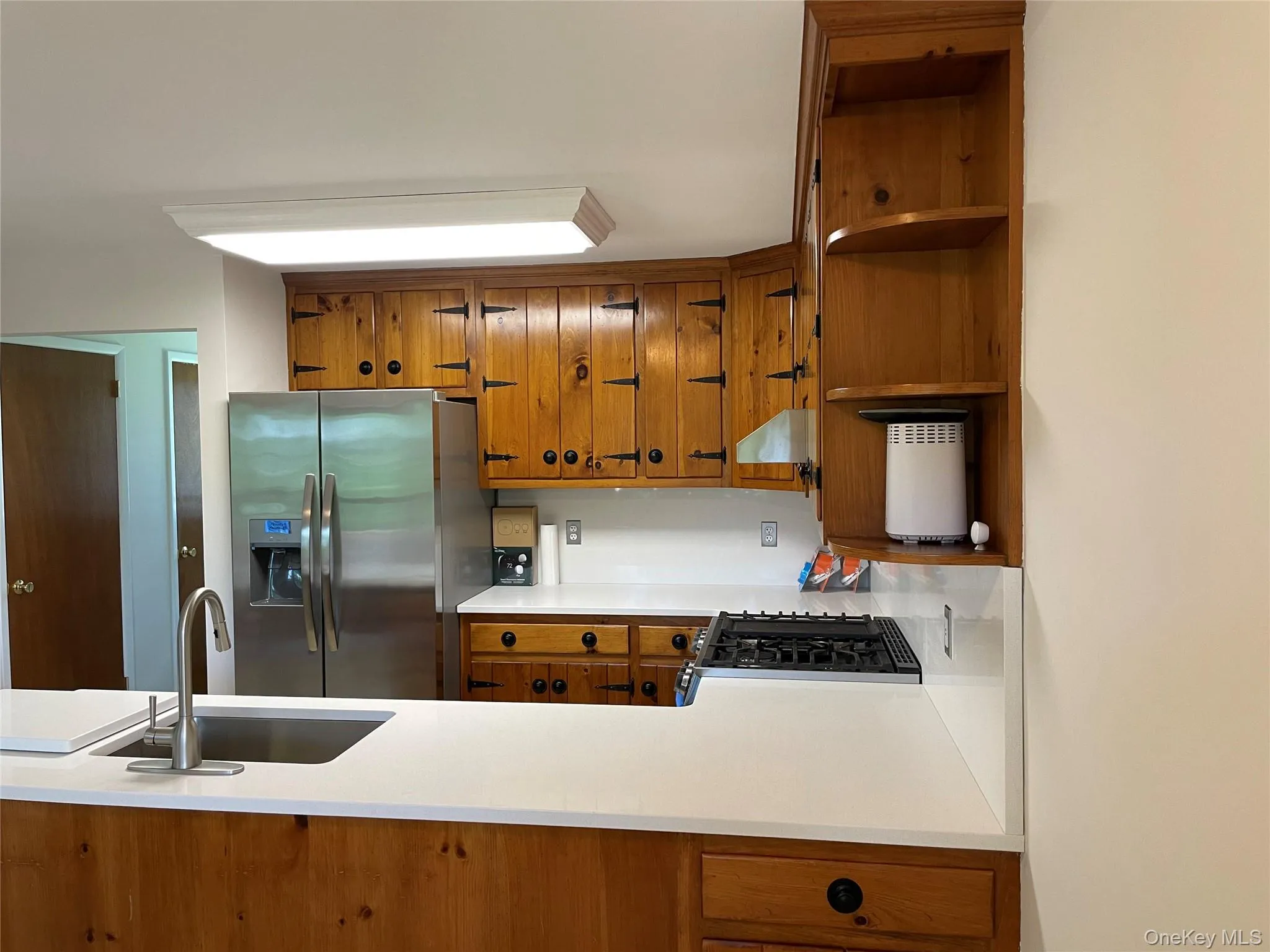 Kitchen featuring stainless steel refrigerator with ice dispenser, brown cabinetry, open shelves, and light countertops Kitchen featuring stainless steel refrigerator with ice dispenser, brown cabinetry, open shelves, and light countertops