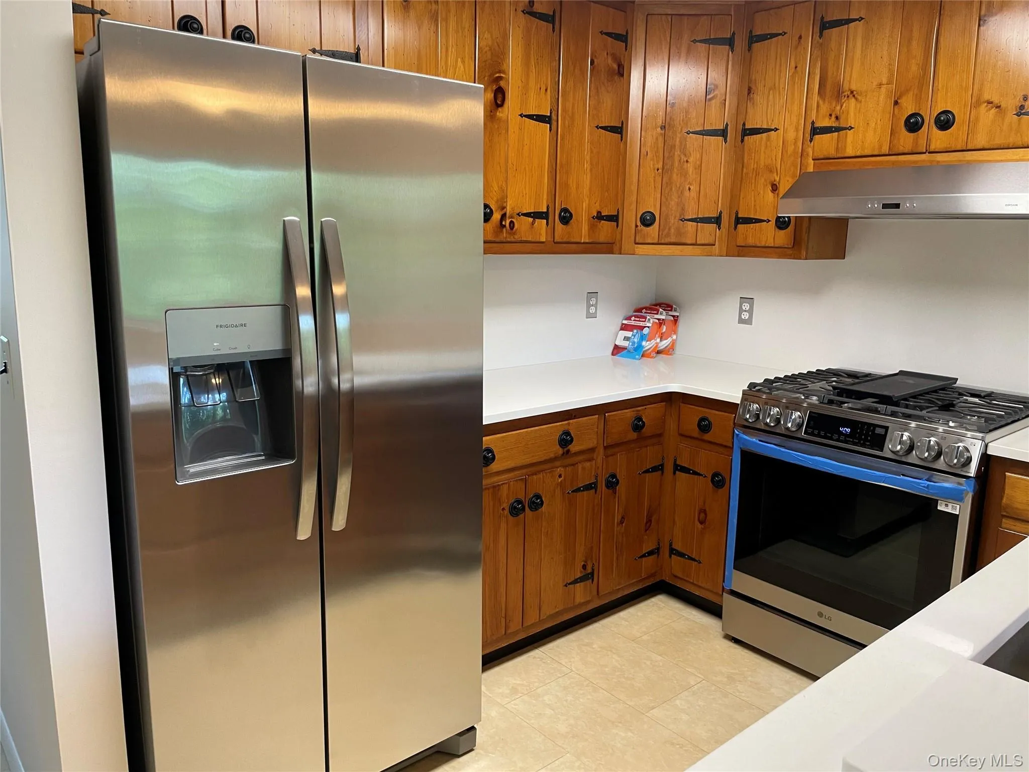 Kitchen featuring stainless steel appliances, under cabinet range hood, brown cabinetry, light countertops, and light tile patterned floors Kitchen featuring stainless steel appliances, under cabinet range hood, brown cabinetry, light countertops, and light tile patterned floors