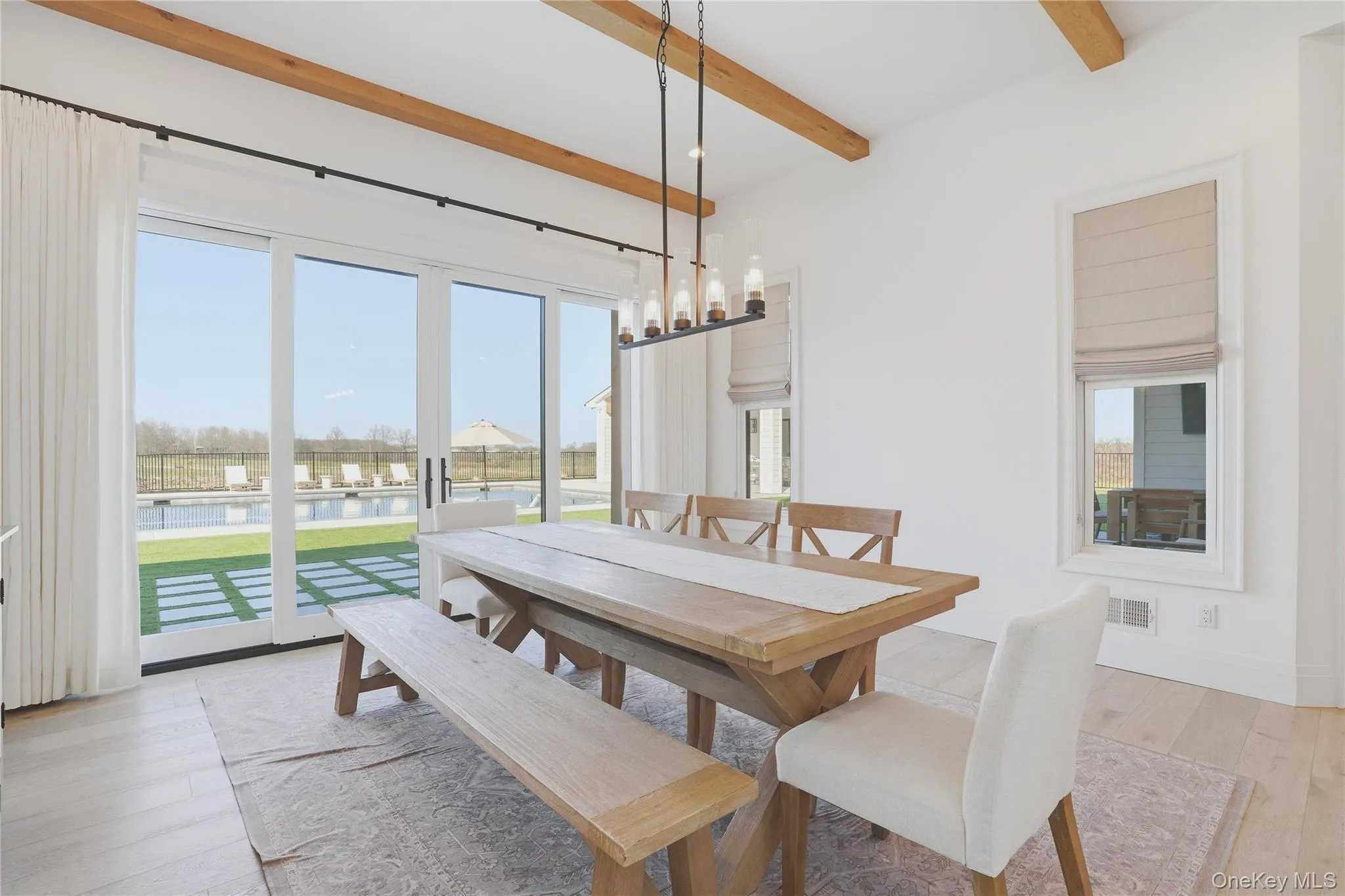 Spacious room featuring exposed wood ceiling beams, light wood-finish flooring, and a contemporary linear chandelier Spacious room featuring exposed wood ceiling beams, light wood-finish flooring, and a contemporary linear chandelier