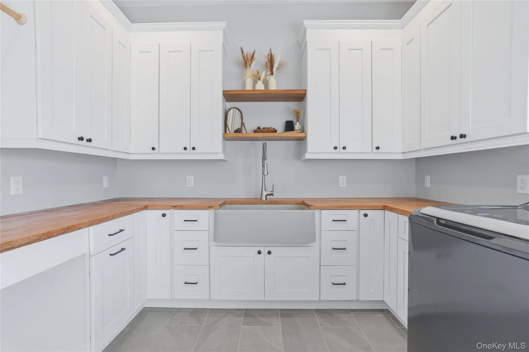 Laundry area featuring extensive white shaker cabinetry, butcher block countertops, a large farmhouse-style sink with a gooseneck faucet, two open wood shelves, and grey tile flooring Laundry area featuring extensive white shaker cabinetry, butcher block countertops, a large farmhouse-style sink with a gooseneck faucet, two open wood shelves, and grey tile flooring