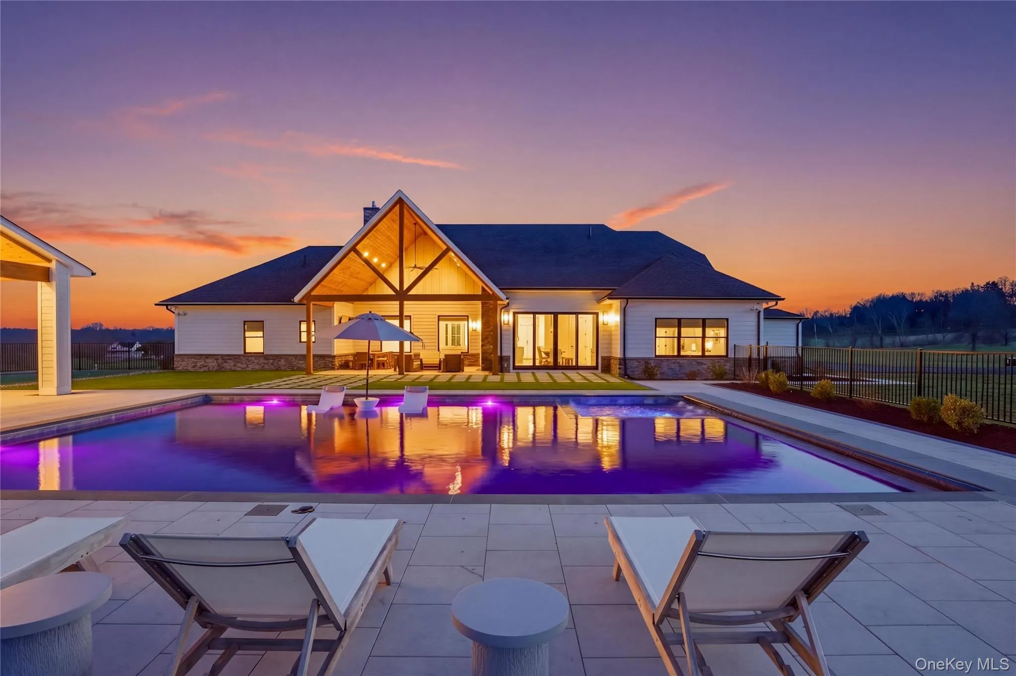 Expansive outdoor living area featuring a rectangular swimming pool with integrated lighting, a spacious patio with paver flooring, a covered outdoor lounge with exposed beams, and a white exterior home with stone accents Expansive outdoor living area featuring a rectangular swimming pool with integrated lighting, a spacious patio with paver flooring, a covered outdoor lounge with exposed beams, and a white exterior home with stone accents