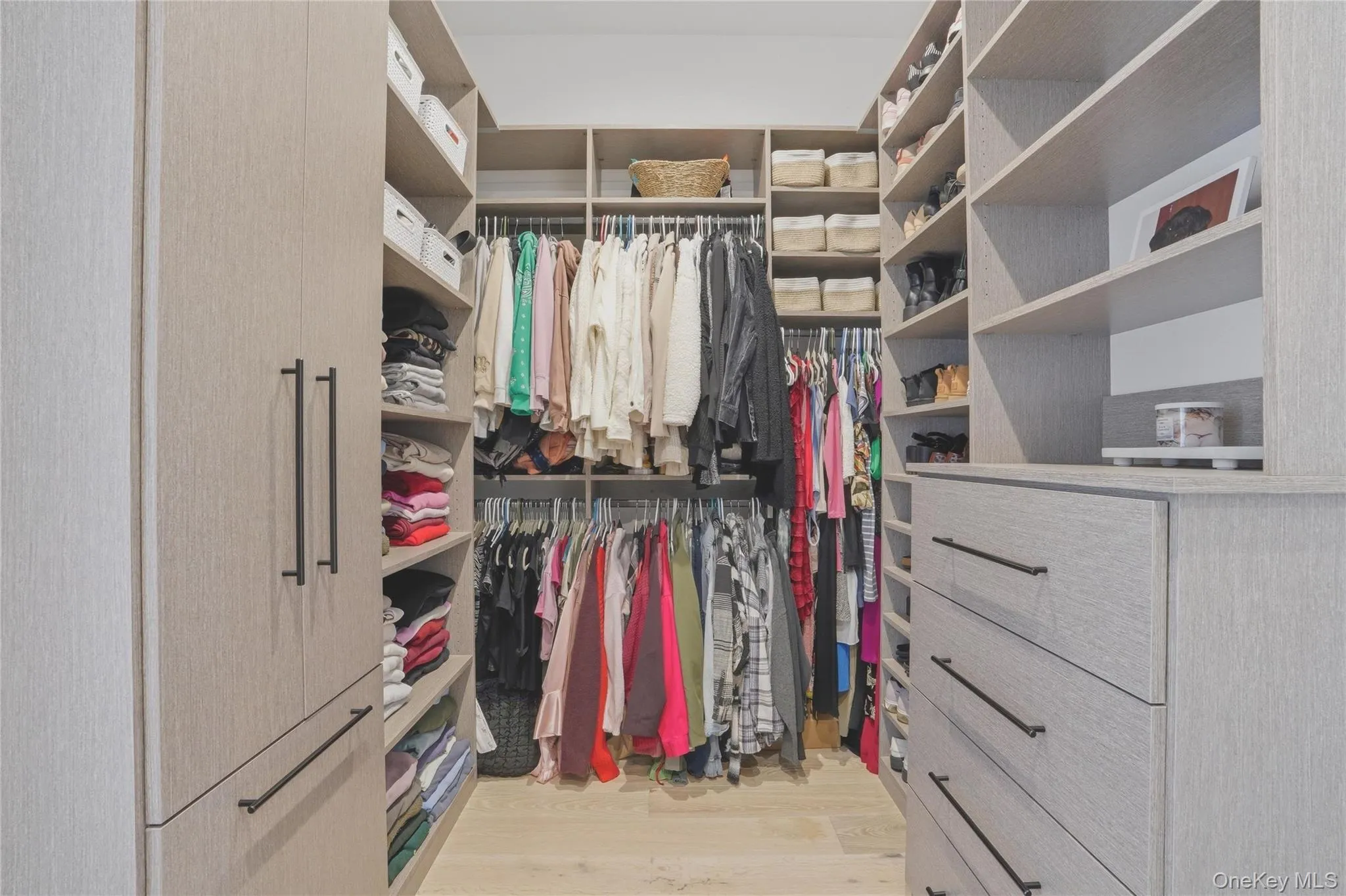 Spacious U-shaped walk-in closet featuring extensive built-in shelving, multiple hanging rods, integrated drawers with linear pulls, and wood-finish flooring Spacious U-shaped walk-in closet featuring extensive built-in shelving, multiple hanging rods, integrated drawers with linear pulls, and wood-finish flooring