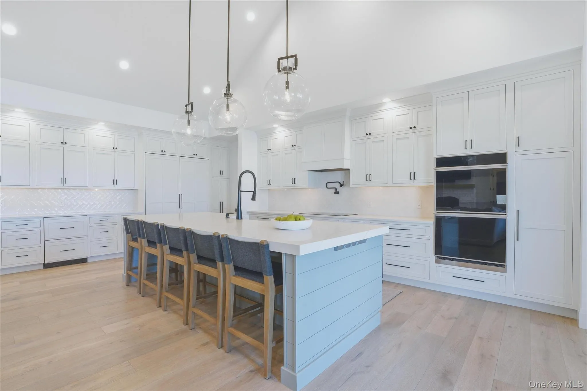 Bright kitchen featuring a large center island with seating, light wood-finish flooring, white cabinetry, integrated double ovens, and a herringbone pattern tile backsplash Bright kitchen featuring a large center island with seating, light wood-finish flooring, white cabinetry, integrated double ovens, and a herringbone pattern tile backsplash