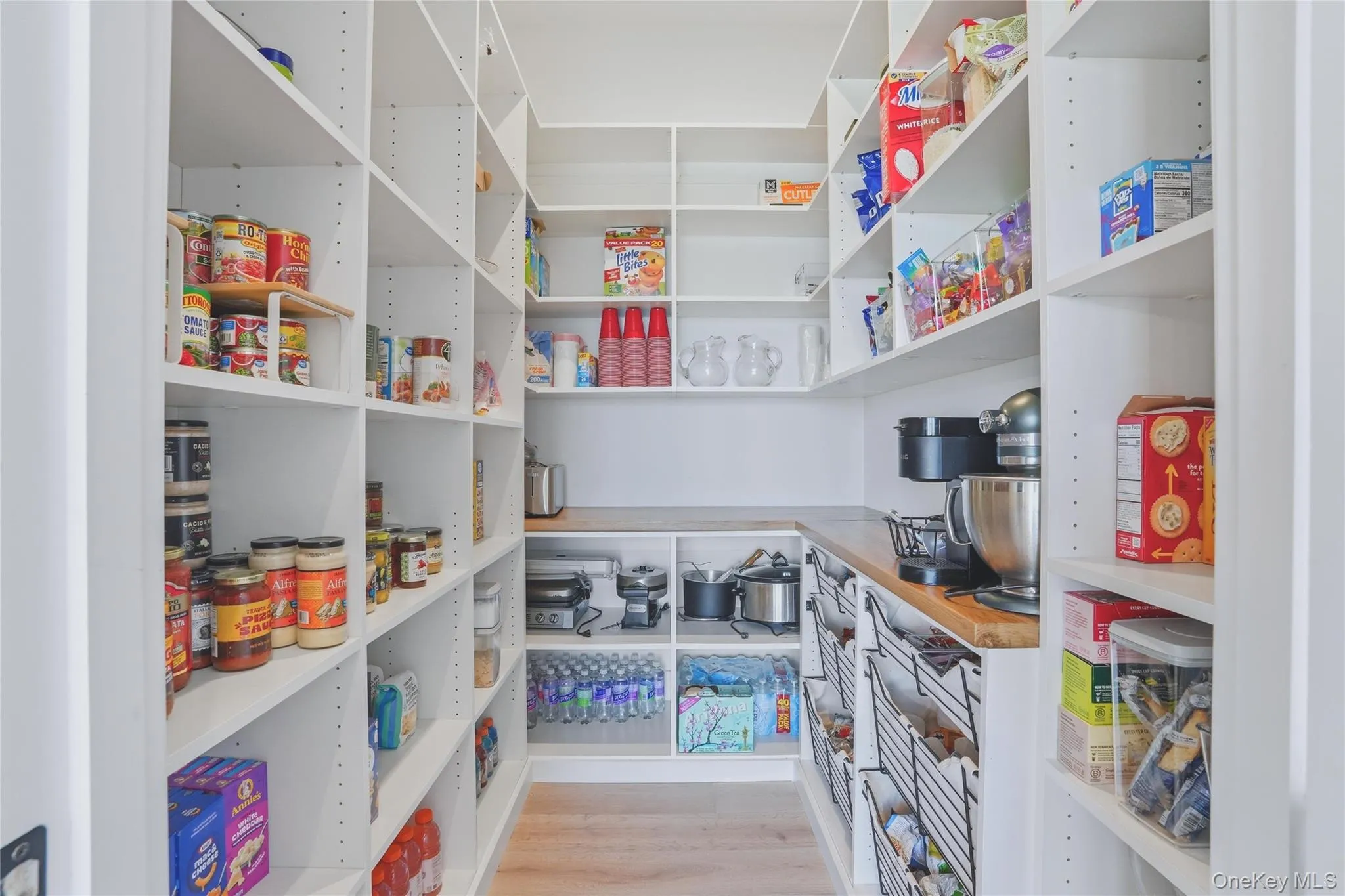 Walk-in pantry featuring extensive white shelving, wood-finish flooring, a wood-finish countertop, under-counter storage, and built-in wire baskets Walk-in pantry featuring extensive white shelving, wood-finish flooring, a wood-finish countertop, under-counter storage, and built-in wire baskets