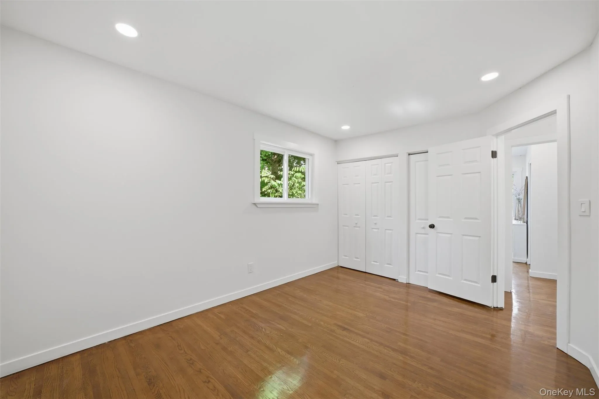 Spacious 2nd main floor bedroom featuring wood-finish flooring, recessed lighting, a window with exterior foliage, and two bi-fold closet doors Spacious 2nd main floor bedroom featuring wood-finish flooring, recessed lighting, a window with exterior foliage, and two bi-fold closet doors