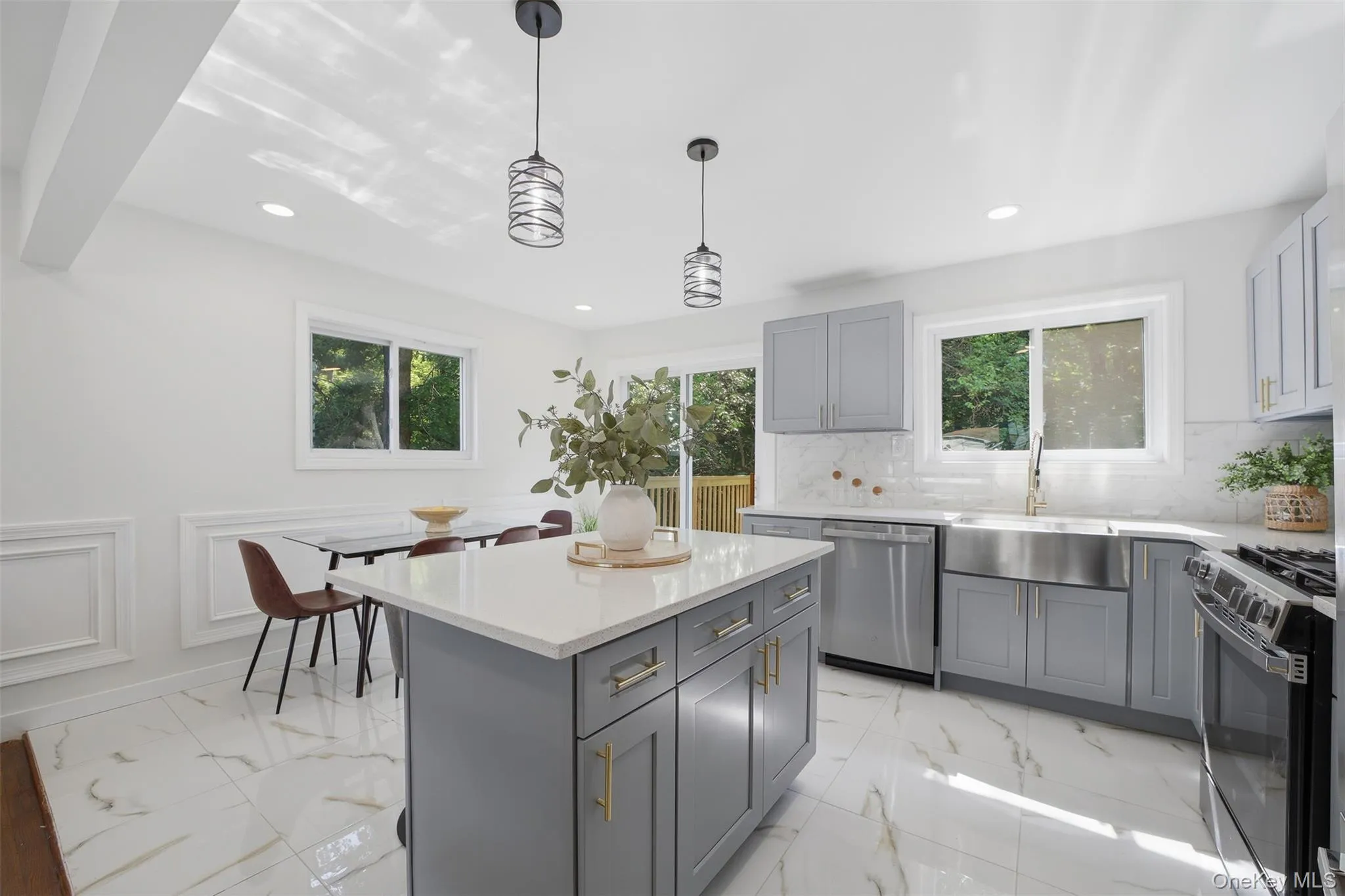 Modern kitchen featuring a central island with a light-toned countertop, gray cabinetry with gold-finish hardware, stainless steel appliances, and white marble-look flooring Modern kitchen featuring a central island with a light-toned countertop, gray cabinetry with gold-finish hardware, stainless steel appliances, and white marble-look flooring