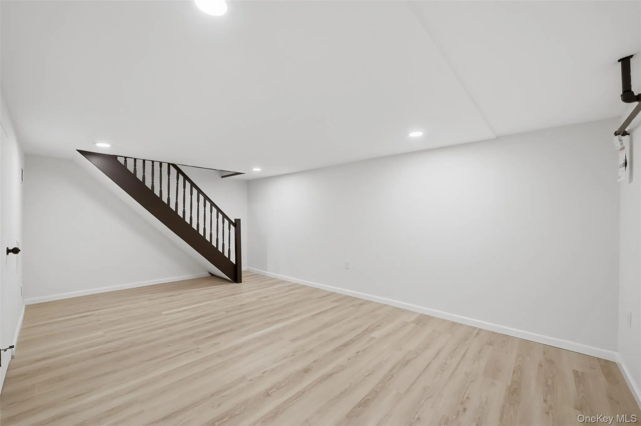 Spacious finished basement featuring light wood-finish flooring, recessed lighting, and a modern dark wood staircase with vertical balusters Spacious finished basement featuring light wood-finish flooring, recessed lighting, and a modern dark wood staircase with vertical balusters