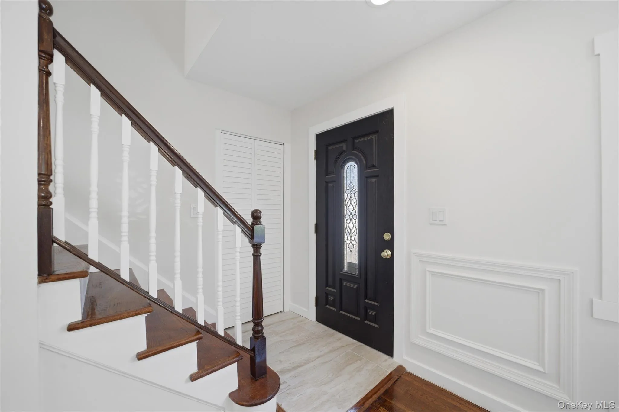 Entryway featuring a dark wood front door with decorative glass inserts, light tile flooring, and white wall paneling Entryway featuring a dark wood front door with decorative glass inserts, light tile flooring, and white wall paneling