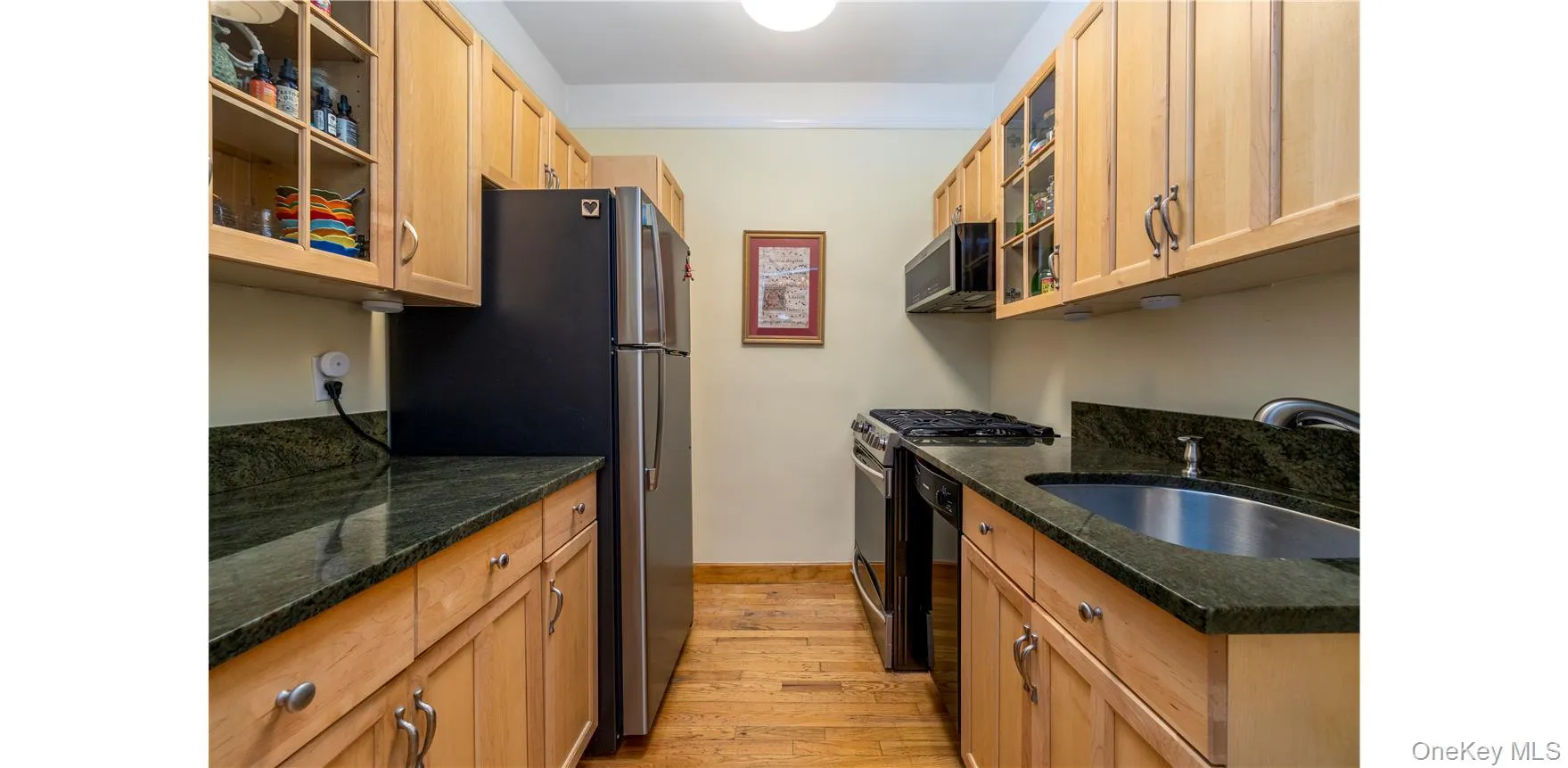 Galley kitchen featuring light wood cabinetry, granite countertops, stainless steel appliances, a built-in microwave, and wood-finish flooring Galley kitchen featuring light wood cabinetry, granite countertops, stainless steel appliances, a built-in microwave, and wood-finish flooring
