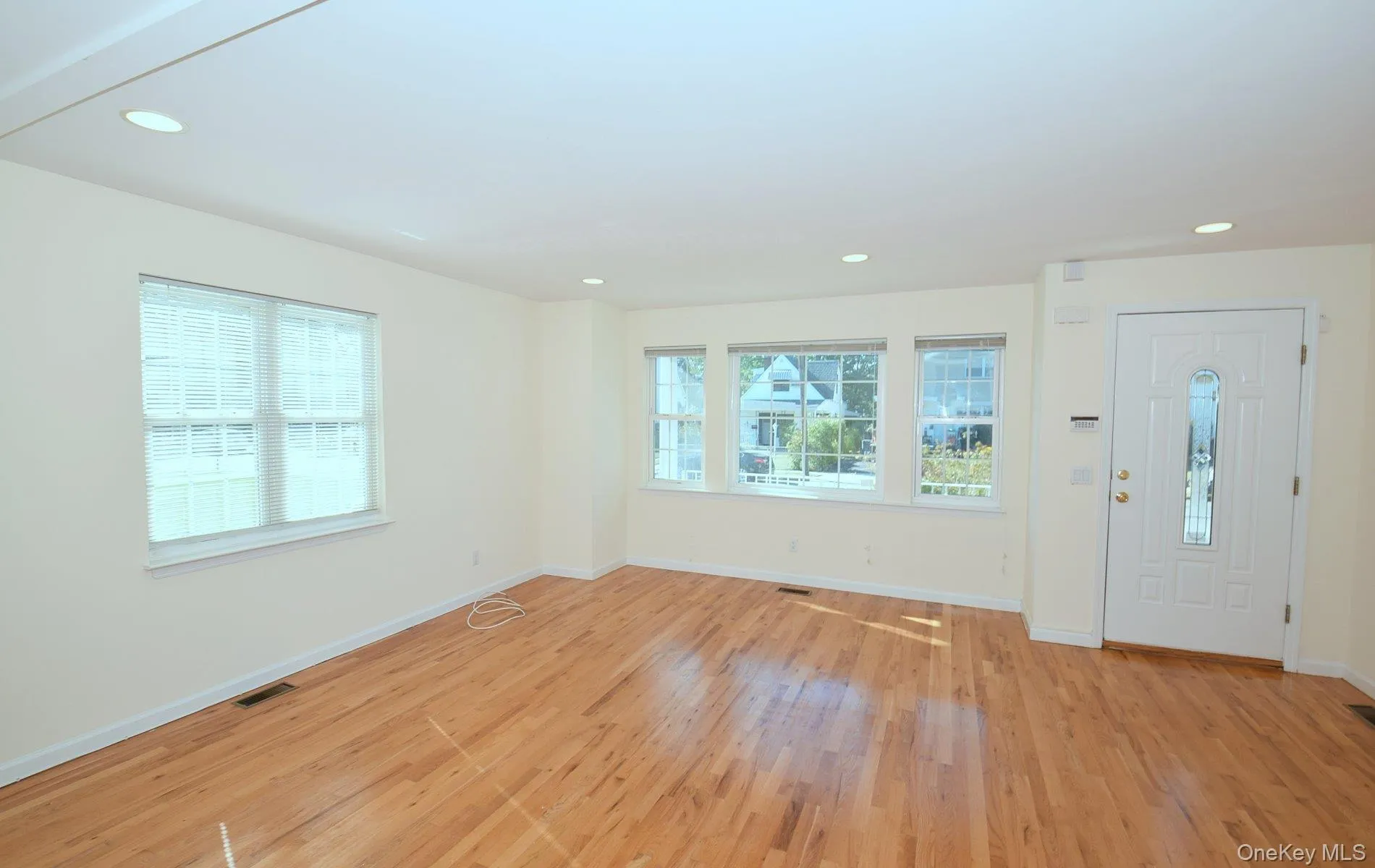 Spacious room featuring wood flooring, recessed lighting, multiple windows with blinds, a white entry door with an arched glass insert, and light-colored wall finishes Spacious room featuring wood flooring, recessed lighting, multiple windows with blinds, a white entry door with an arched glass insert, and light-colored wall finishes