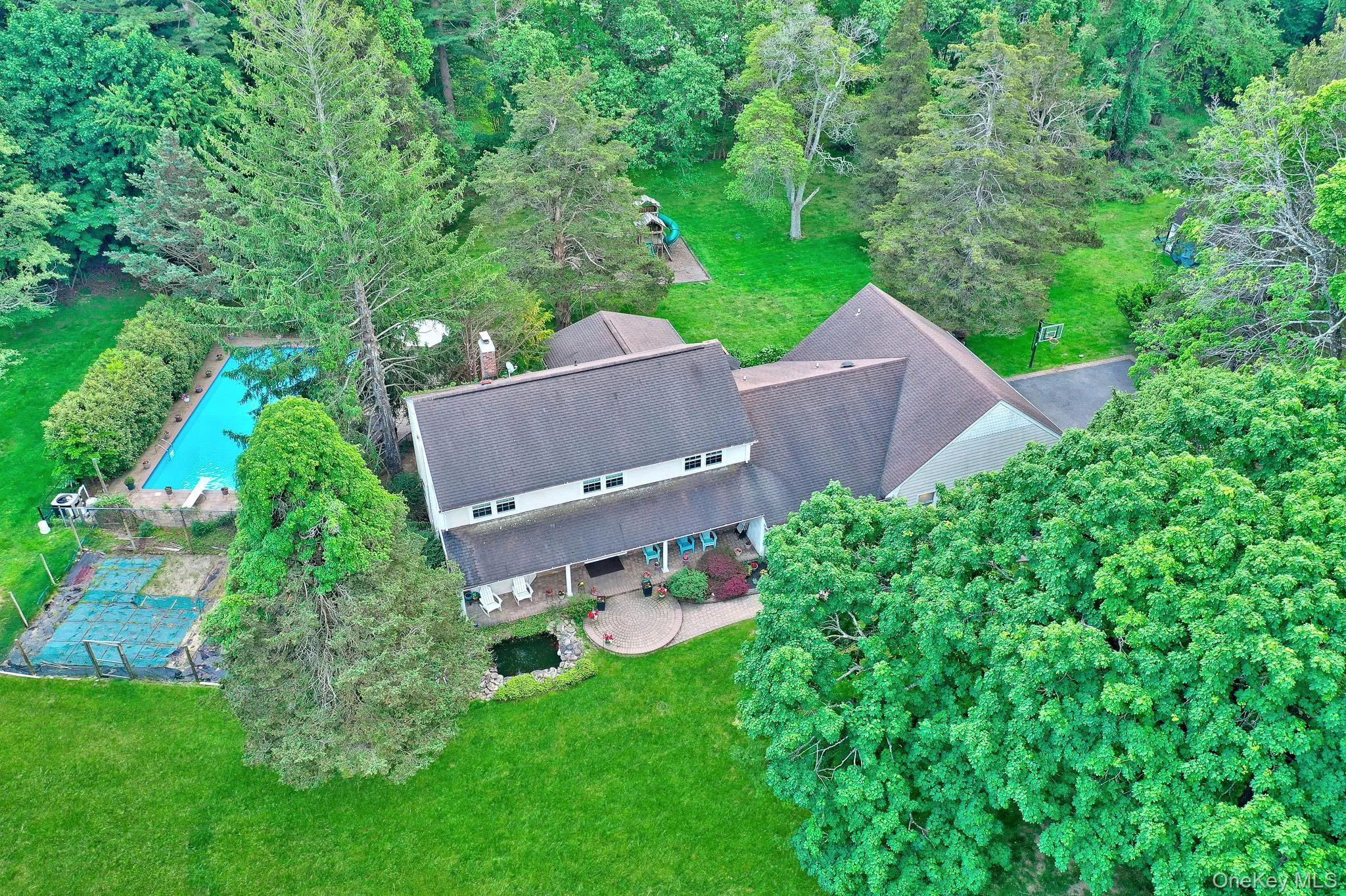 Expansive property featuring a multi-level residence with a gray shingle roof, a rectangular in-ground swimming pool, a dedicated sport court, a stone patio, and mature tree landscaping Expansive property featuring a multi-level residence with a gray shingle roof, a rectangular in-ground swimming pool, a dedicated sport court, a stone patio, and mature tree landscaping