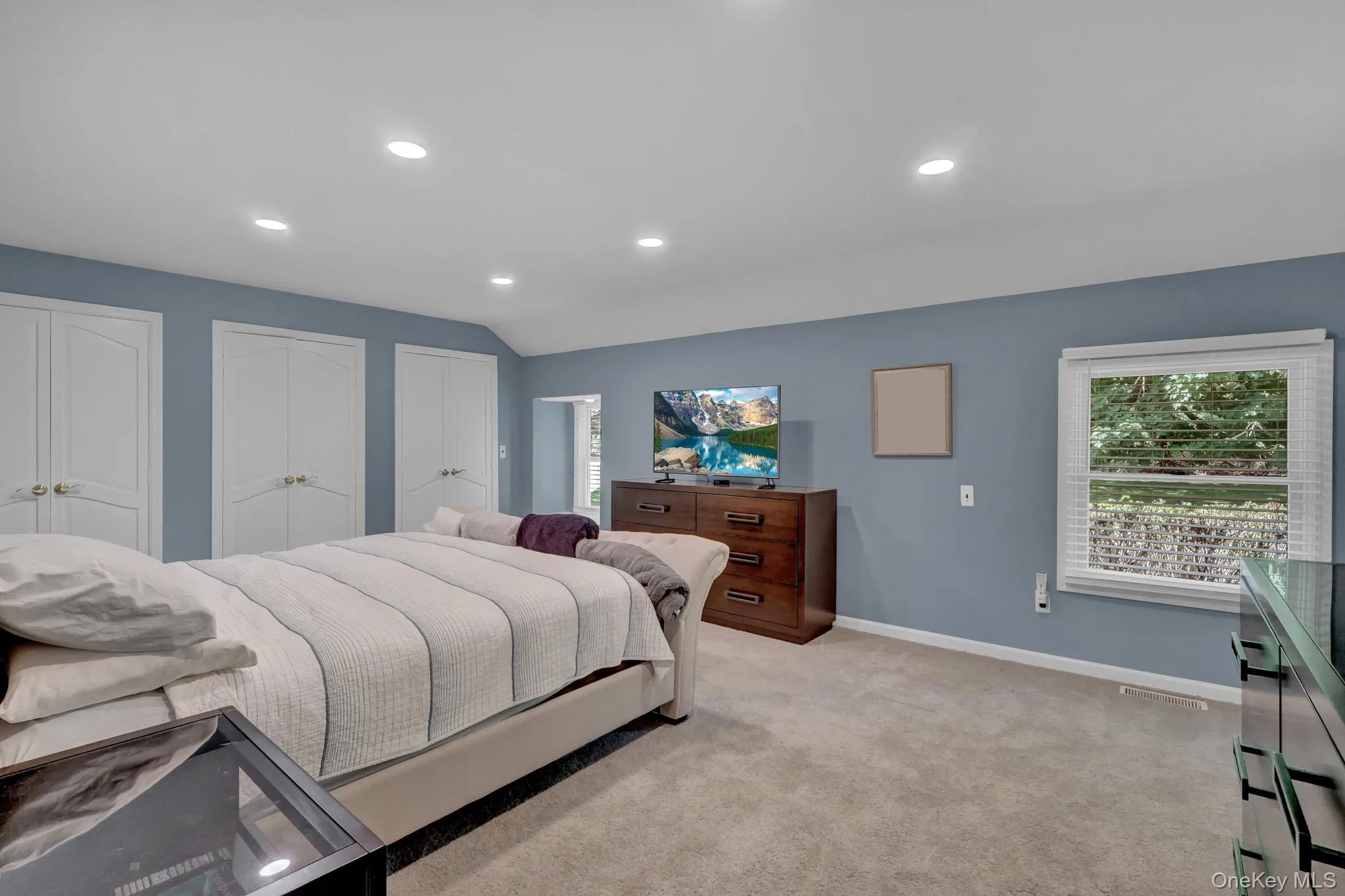 Spacious room featuring light blue walls, recessed lighting, carpet flooring, a window with horizontal blinds, and three sets of white bi-fold closet doors Spacious room featuring light blue walls, recessed lighting, carpet flooring, a window with horizontal blinds, and three sets of white bi-fold closet doors