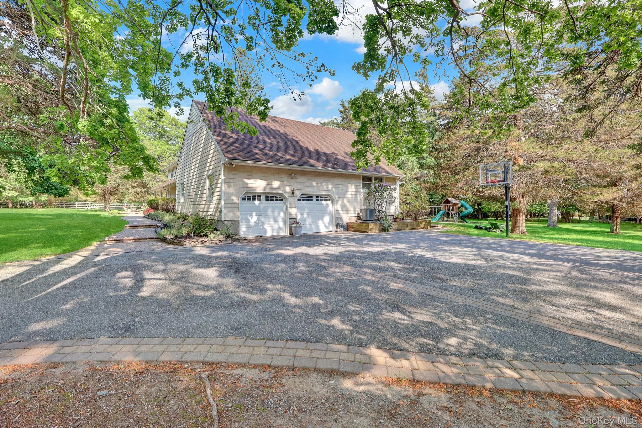 Spacious asphalt driveway featuring a paver stone border, an attached two-car garage with white paneled doors, and mature trees providing natural shade Spacious asphalt driveway featuring a paver stone border, an attached two-car garage with white paneled doors, and mature trees providing natural shade