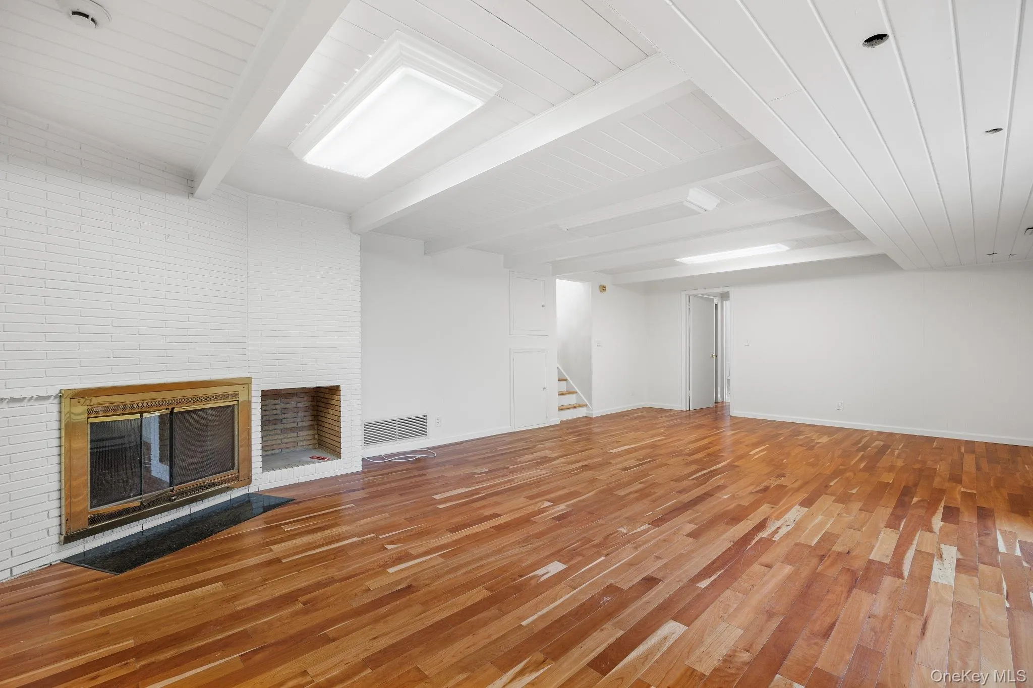 Unfurnished living room with beam ceiling, a brick fireplace, light wood-type flooring, and stairway Unfurnished living room with beam ceiling, a brick fireplace, light wood-type flooring, and stairway