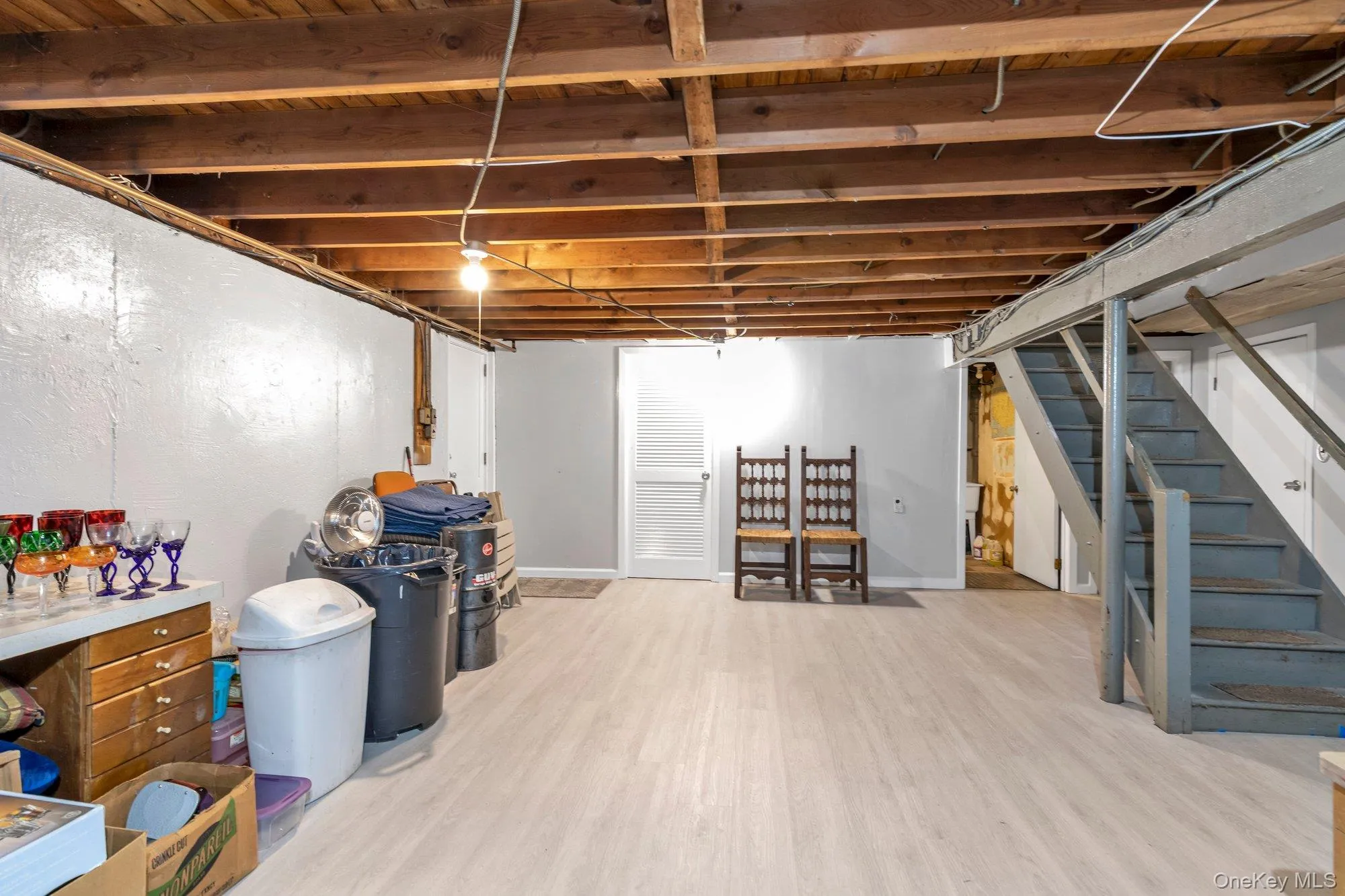 Basement space featuring exposed wood beam ceiling, light laminate-finish flooring, painted concrete walls, and painted wooden stairs Basement space featuring exposed wood beam ceiling, light laminate-finish flooring, painted concrete walls, and painted wooden stairs